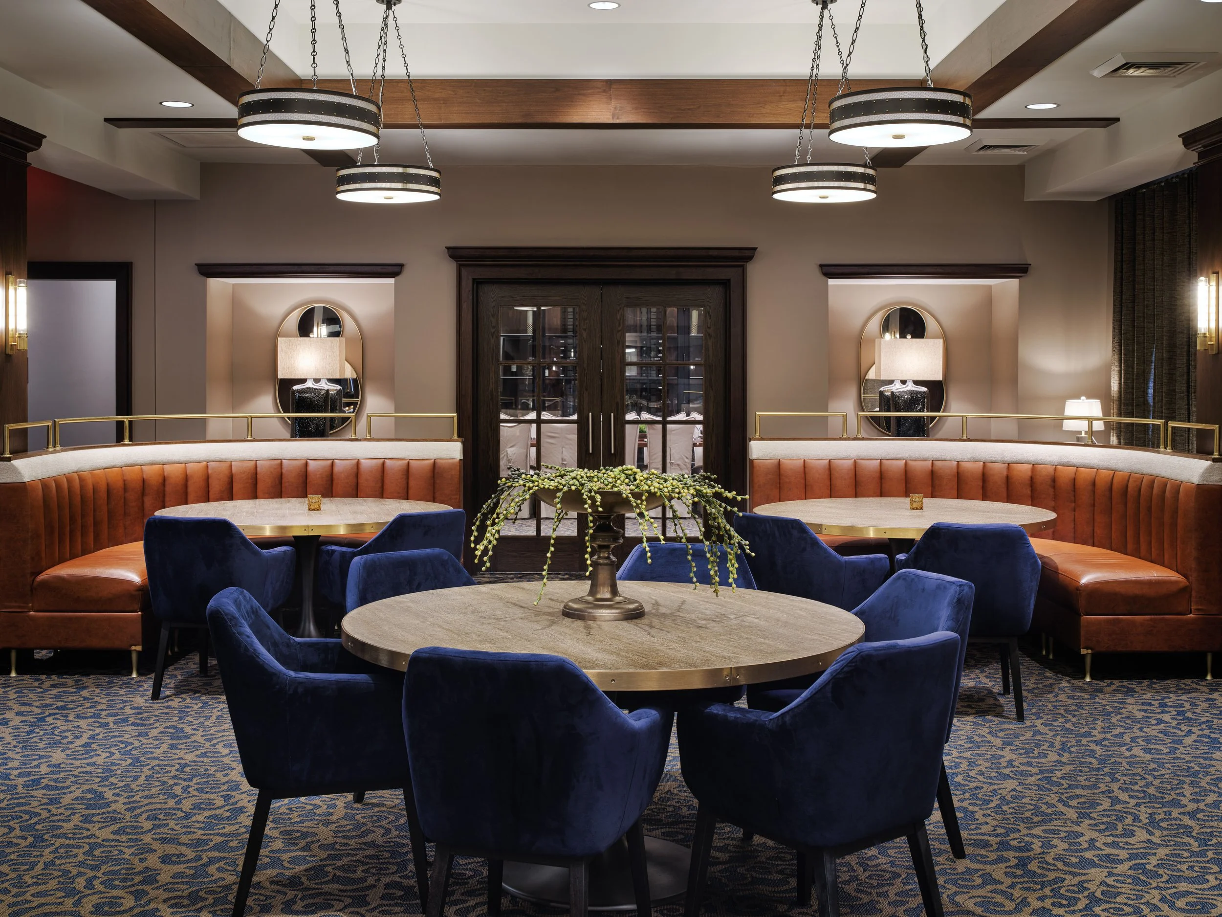 Curved cognac leather banquettes with navy velvet dining chairs, brass-edged tables, and arched mirror niches at Off The Rails, designed by Ashley Schneider