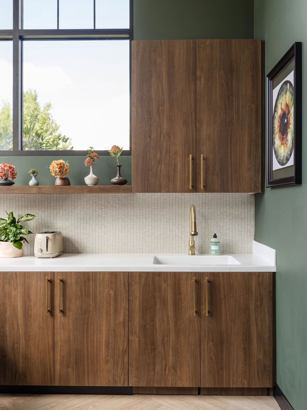 Staff kitchen detail with flat-front walnut cabinets, brass hardware and faucet, white mosaic tile backsplash, and forest green walls with window ledge vase display, interior design by Luxe Style Studio