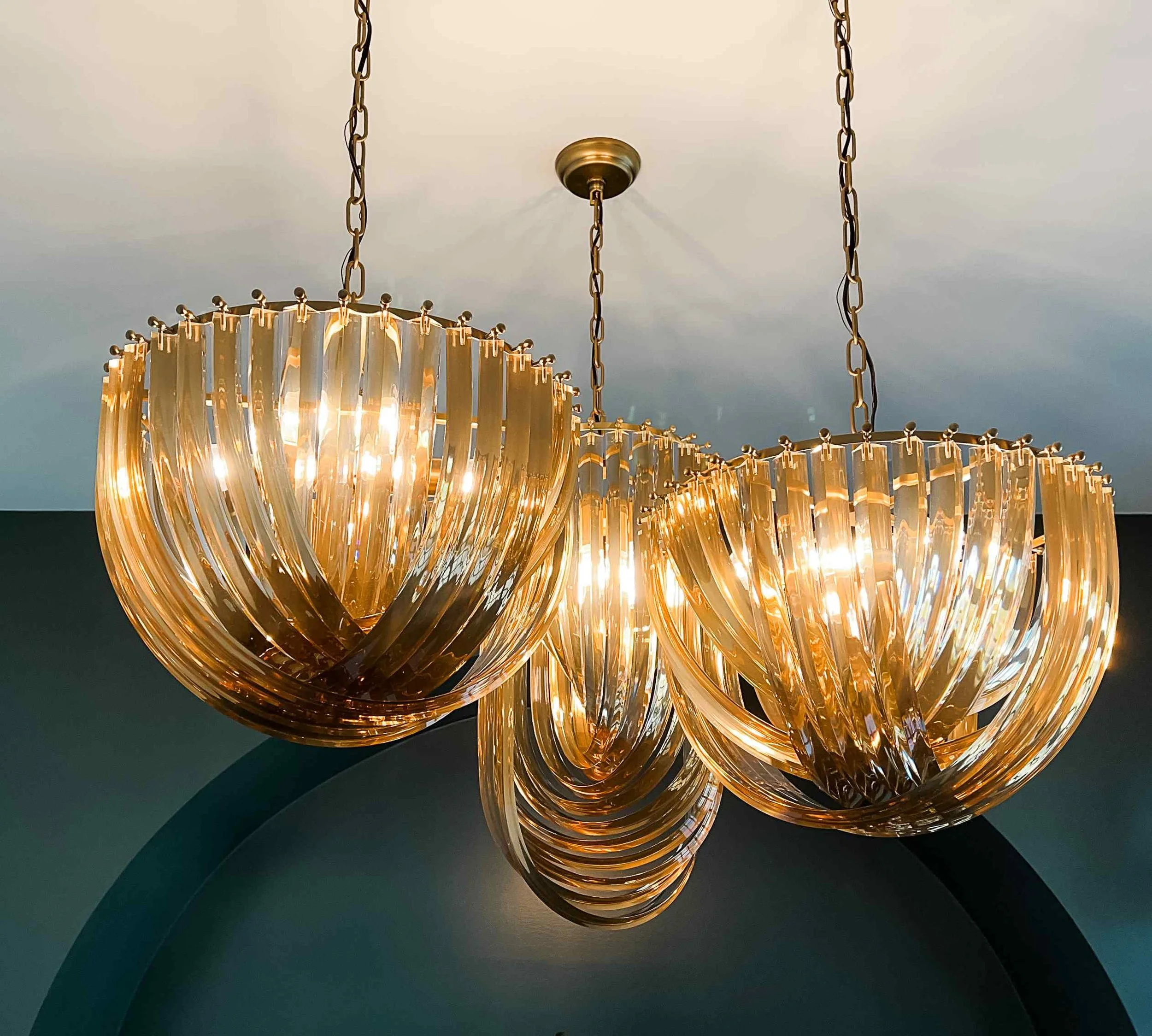 Pair of amber curved-glass Murano-style chandelier pendants against deep teal ceiling, Luxe Style Studio