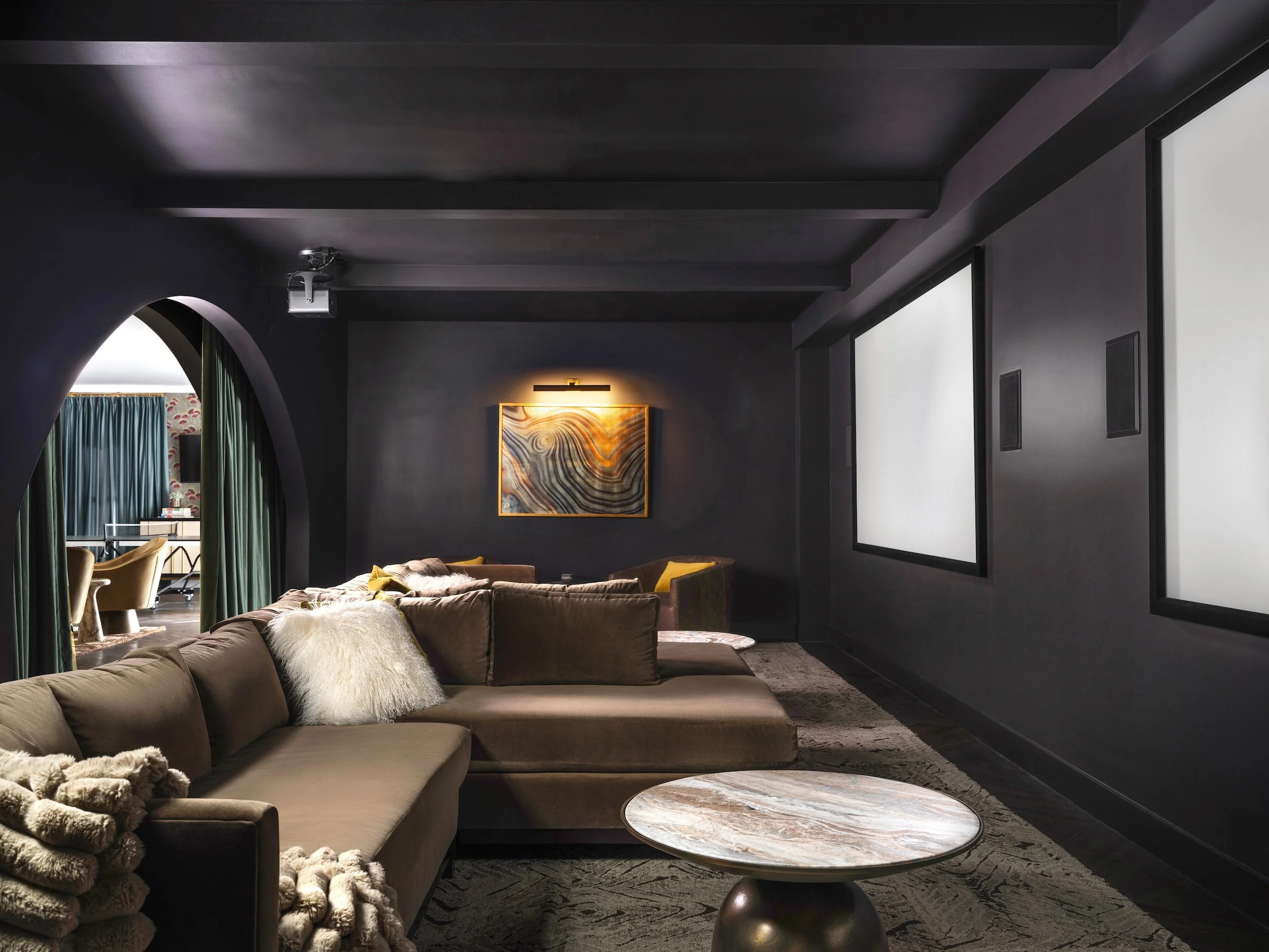 Luxury home theater with dark charcoal walls and ceiling beams, oversized velvet sectional, onyx side table, arched doorway, and dual large projection screens, Atlanta, GA private residence, interior design by Luxe Style Studio