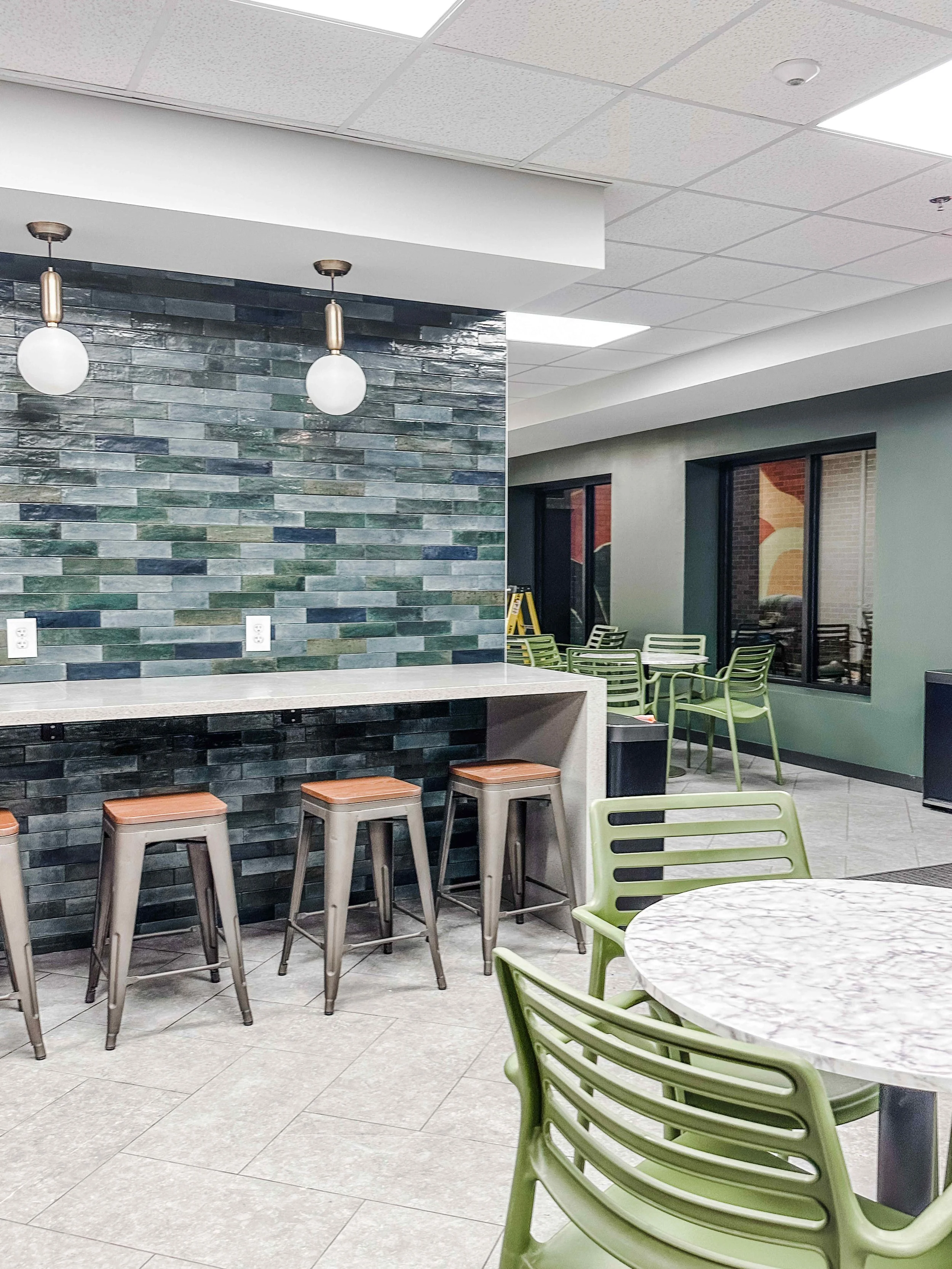 Corporate break room wide view with zellige tile accent wall, white quartz bar counter with metal stools, sage green cafe chairs, round marble bistro tables, and brass globe pendants, interior design by Luxe Style Studio