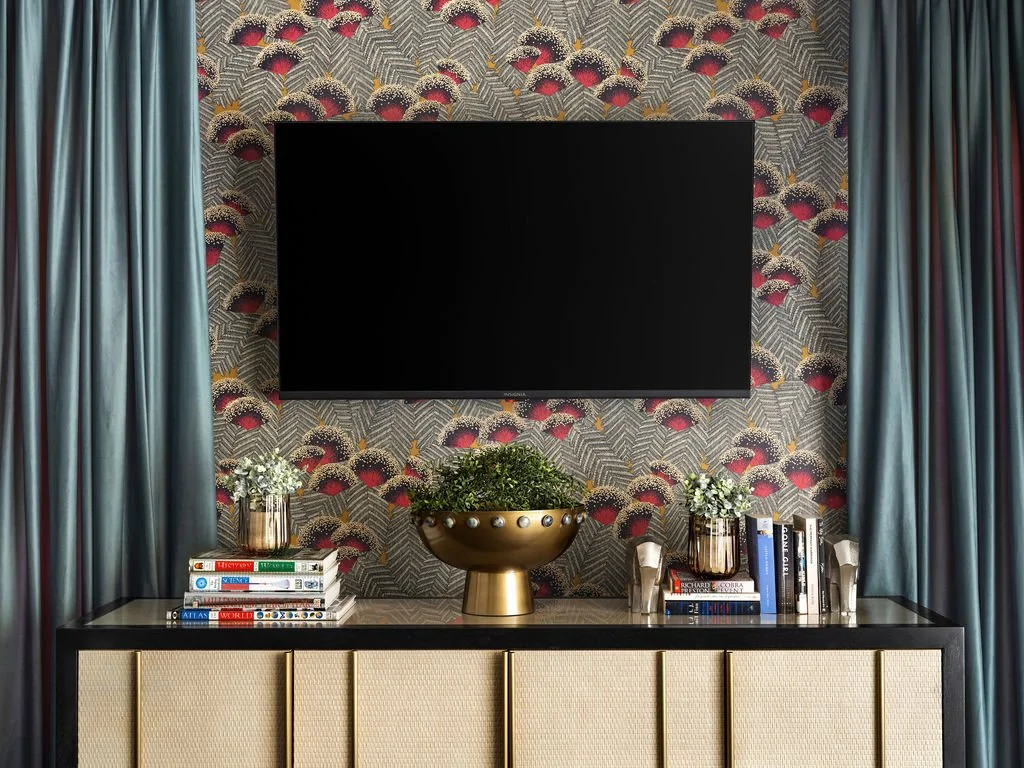 Eclectic TV wall with bold peacock feather wallpaper, cane and brass sideboard styled with books and gold bowl, flanked by teal velvet drapes, interior design by Luxe Style Studio