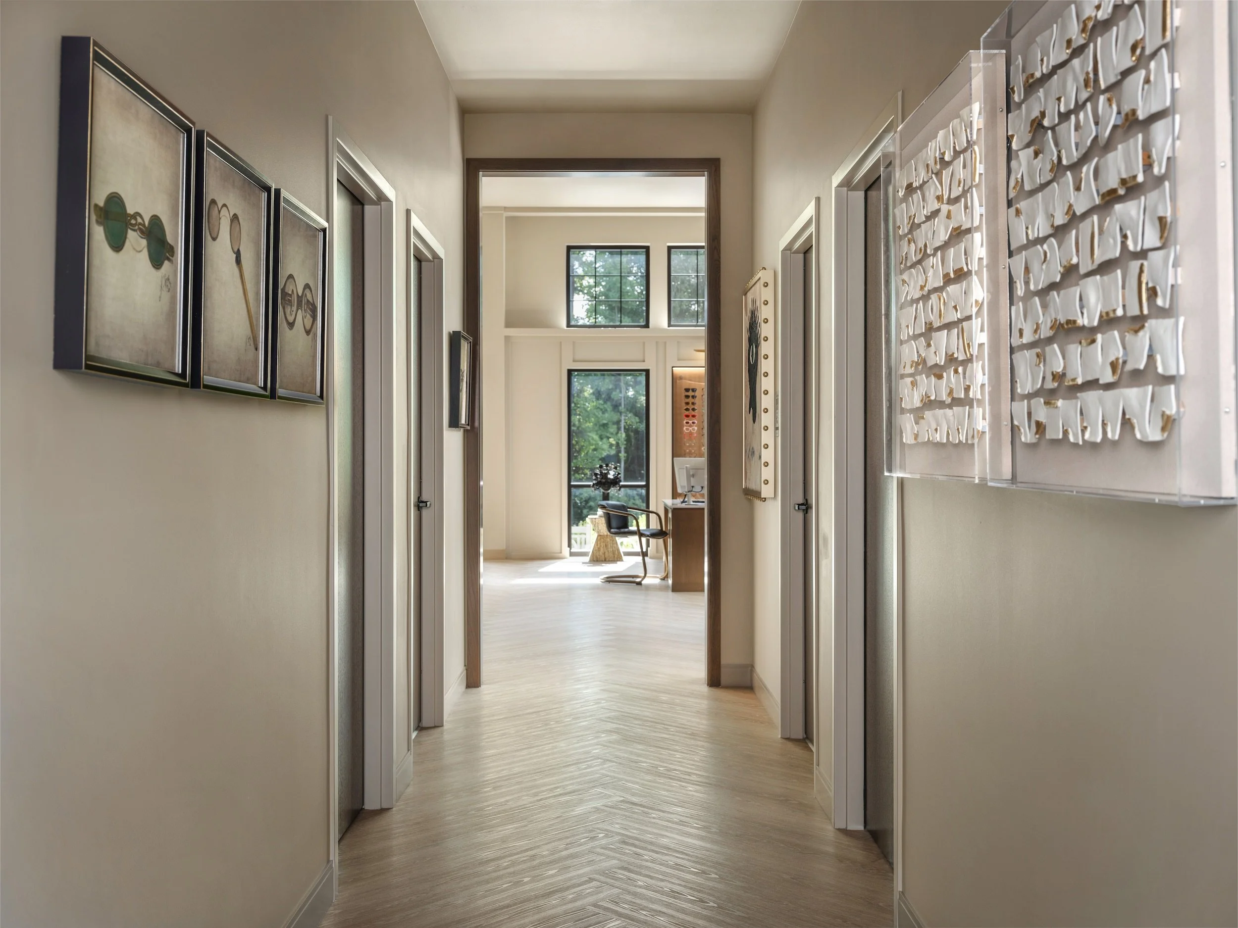 Bright clinical hallway with white oak herringbone floors, framed vintage eyewear artwork, and sculptural wall installation leading to exam rooms, Windsor Eye Care optical boutique, Atlanta, GA, interior design by Luxe Style Studio