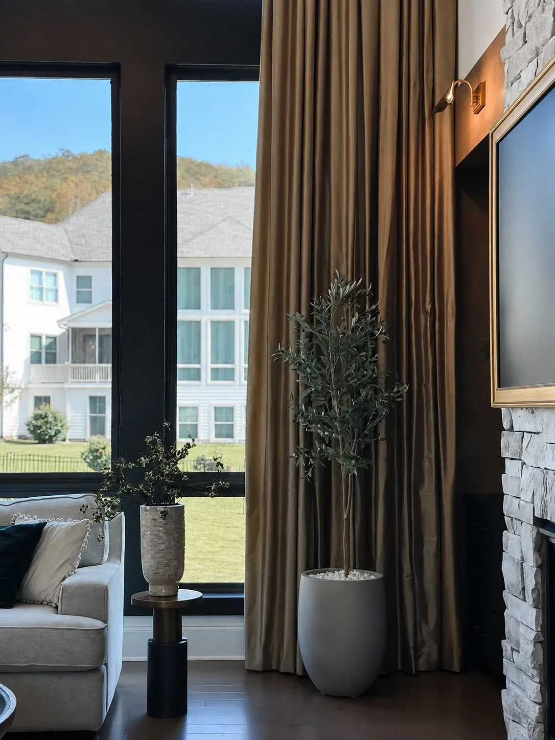 Floor-to-ceiling bronze drapery beside the tall black-framed windows and stone fireplace in the East Cobb great room, designed by Luxe Style Studio
