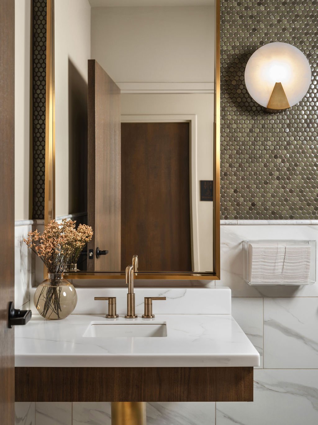 Luxury commercial powder room with metallic penny tile accent wall, brass-framed vanity mirror, globe sconce, brushed brass faucet, and white marble countertop and tile, interior design by Luxe Style Studio