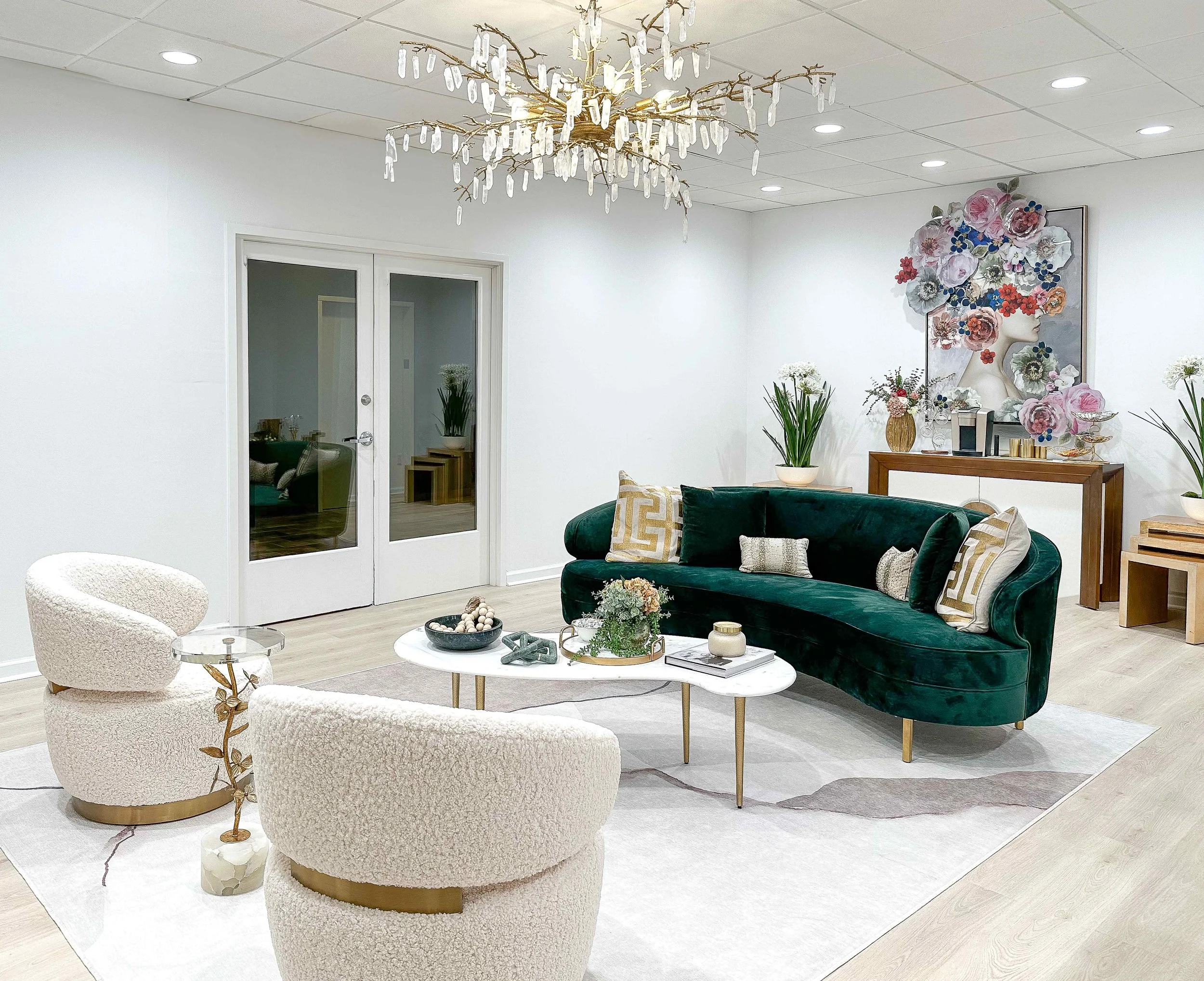 Full view of luxury medical spa waiting room with curved emerald green velvet sofa, cream bouclé chairs, white marble coffee table, branch crystal chandelier, and large floral portrait artwork, interior design by Luxe Style Studio