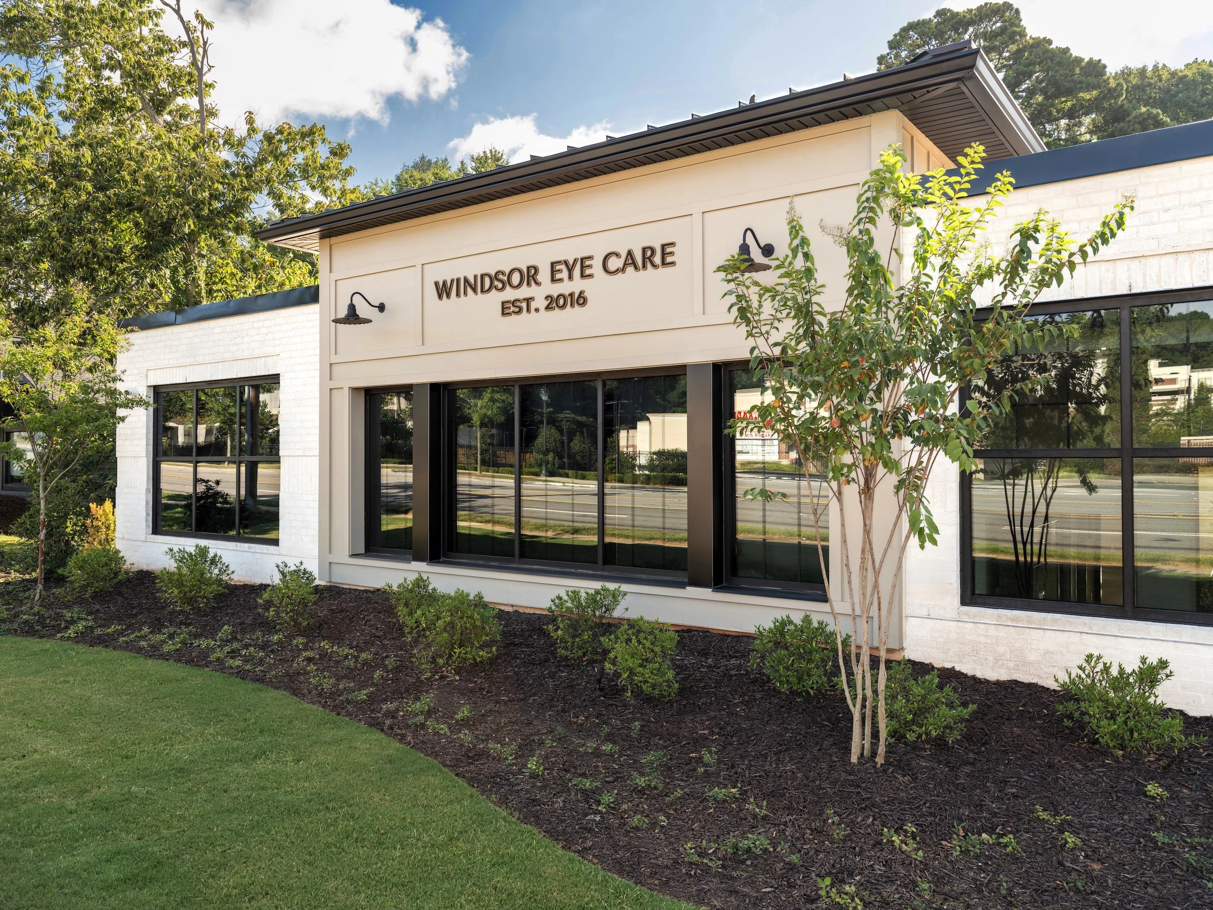 Close-up of white painted brick office building exterior with black steel casement windows and gooseneck sconce lighting, Windsor Eye Care optical boutique, Atlanta, GA, interior design by Luxe Style Studio