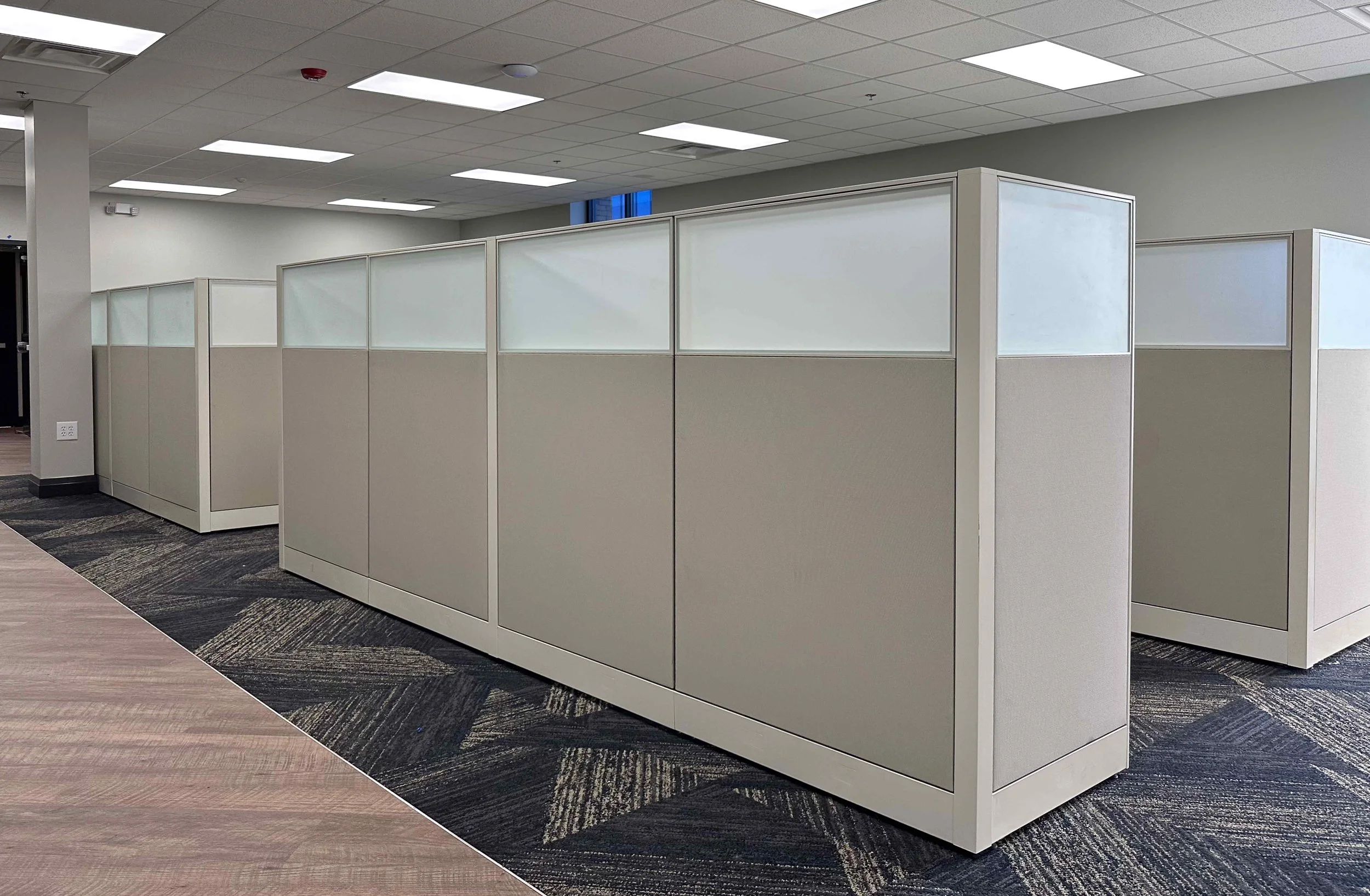 Open plan corporate office with rows of neutral cubicle workstations featuring frosted glass upper panels, geometric patterned carpet tile, and wood plank flooring border, interior design by Luxe Style Studio