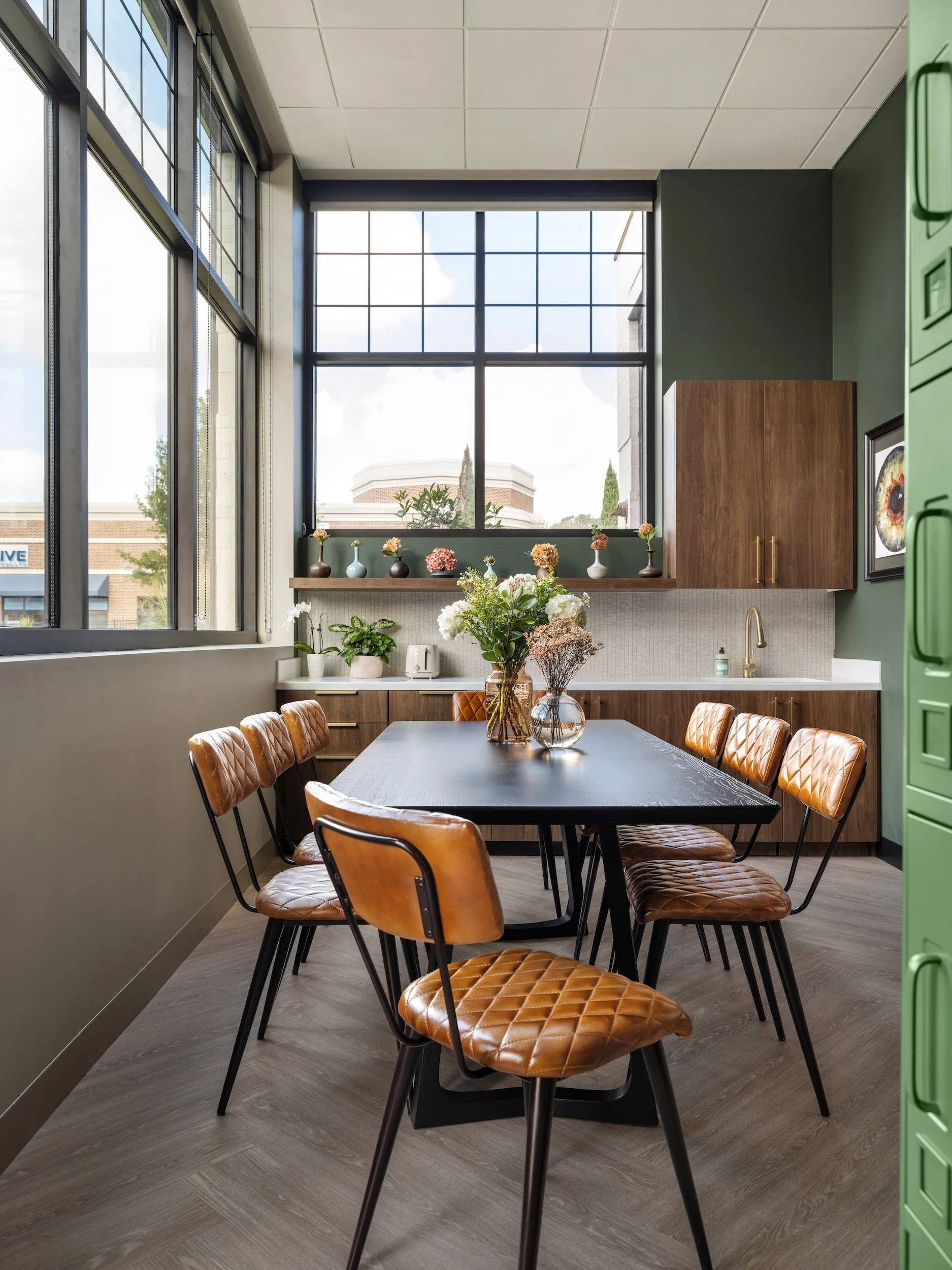 Staff break room with forest green walls, walnut cabinetry, cognac leather chairs, and black-framed windows, designed by Luxe Style Studio
