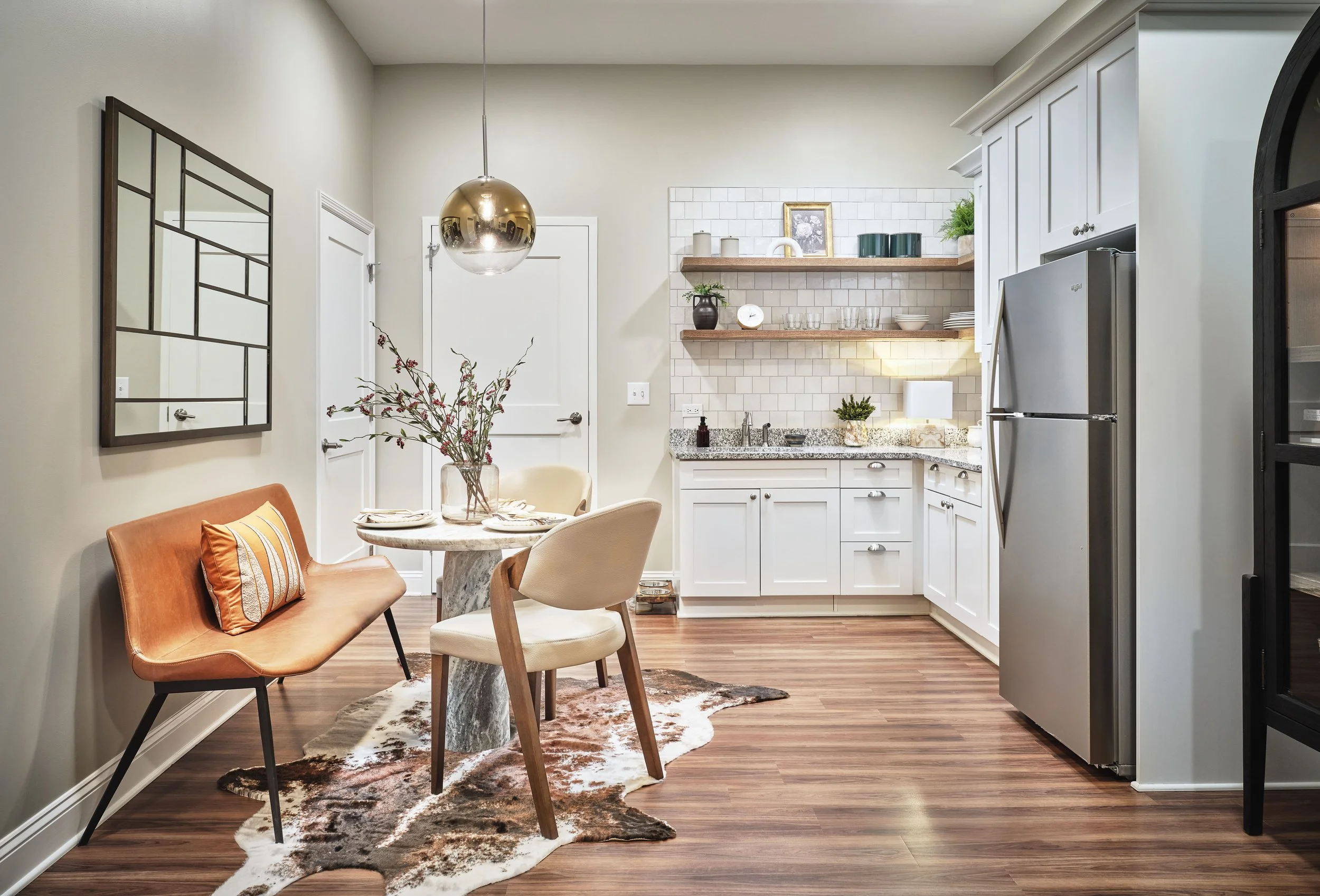 West Broad kitchen and dining area in Sugar Hill with a marble pedestal table, tan leather bench, cream dining chairs, cowhide rug, brass globe pendant, white Shaker cabinets, zellige tile, and open wood shelving, designed by Ashley Schneider