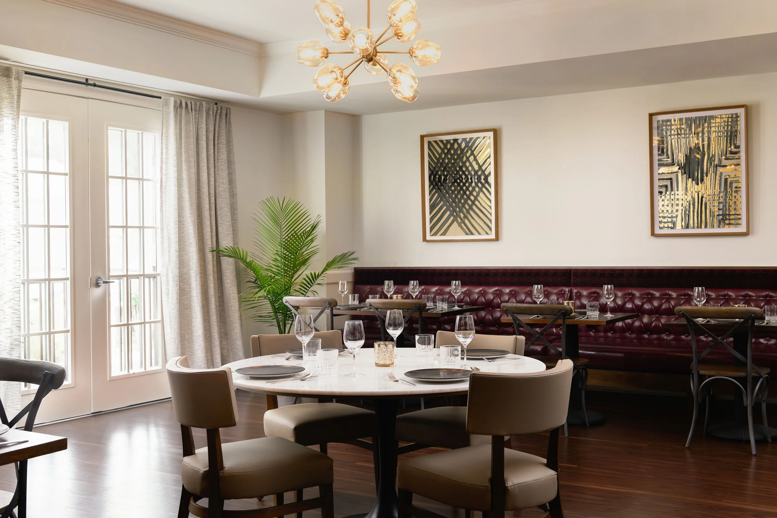 Local Hearth private dining room in Acworth with a burgundy tufted banquette, round marble table, upholstered chairs, brass sputnik chandelier, gold-framed artwork, and French doors, designed by Ashley Schneider