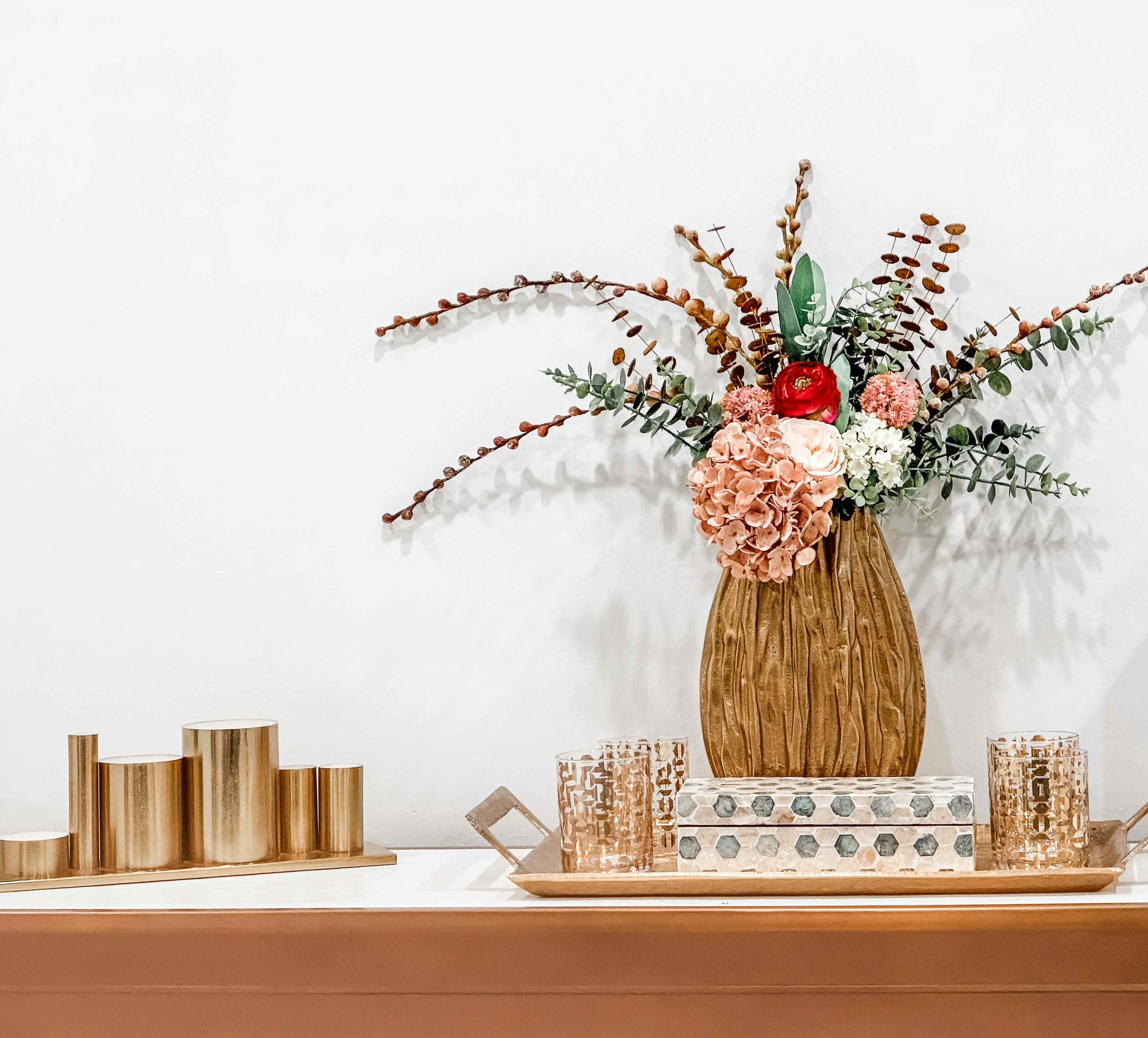 Styled console detail in luxury medical spa with textured gold vase, pink hydrangea floral arrangement, brass candleholders, and gold tray with crystal glasses, interior design by Luxe Style Studio