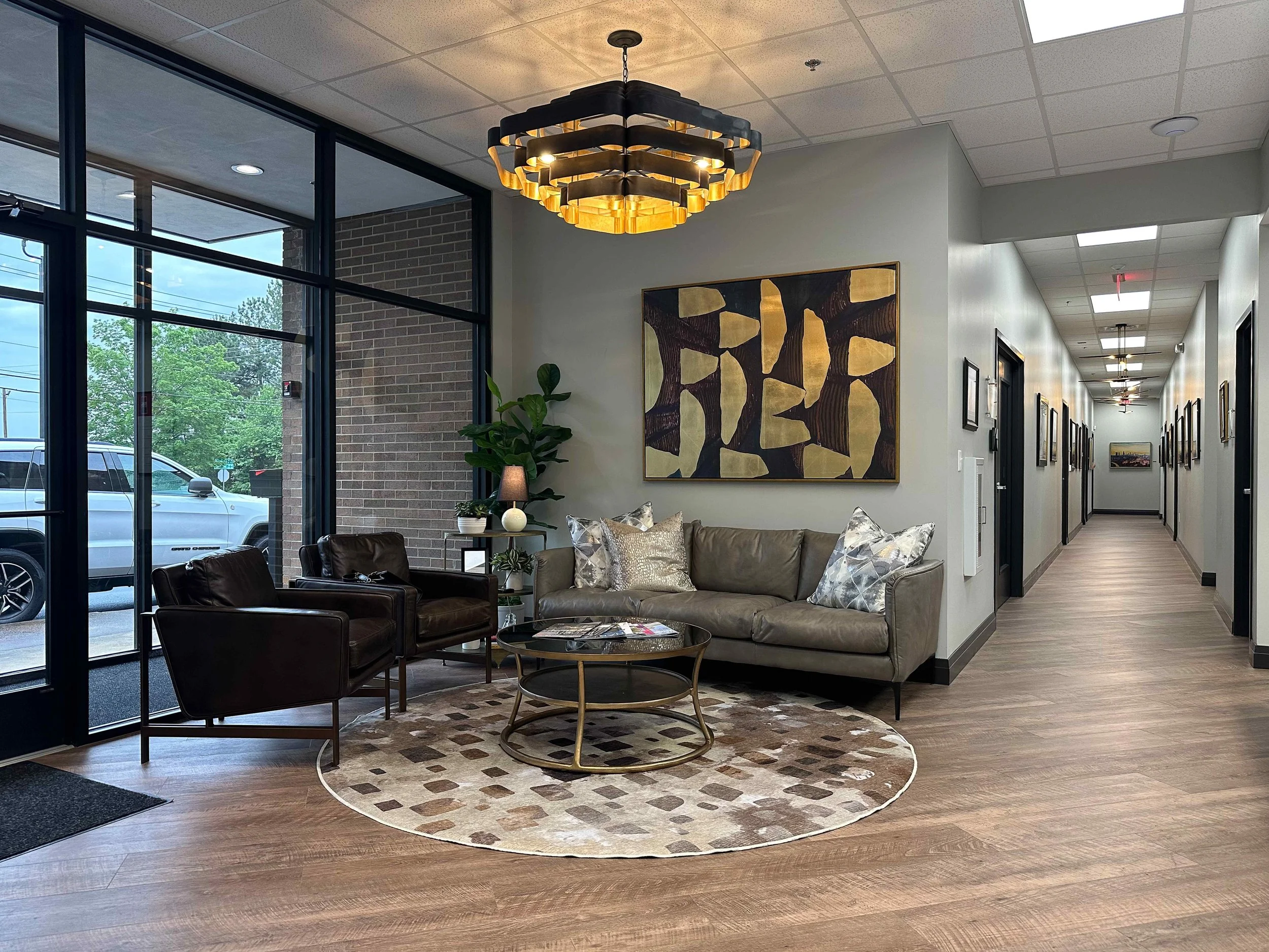 Full view of corporate office reception lobby with taupe leather sofa, dark leather club chairs, cowhide patchwork rug, glass and brass coffee table, gold leaf abstract art, dramatic layered brass chandelier, and gallery hallway beyond with black ste