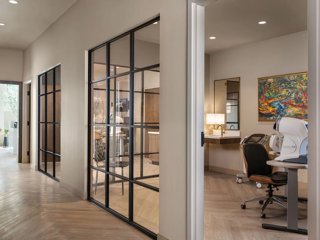 Luxury optometry office corridor with black steel and glass partitions revealing exam rooms, herringbone wood floors, and natural light, interior design by Luxe Style Studio
