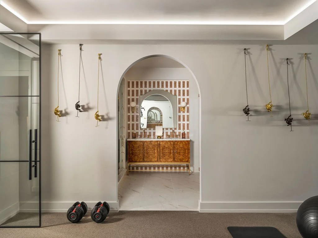 Luxury home gym wall with suspended climbing figure sculptures in gold and bronze, LED cove ceiling lighting, and arched entry to tiled bathroom, interior design by Luxe Style Studio