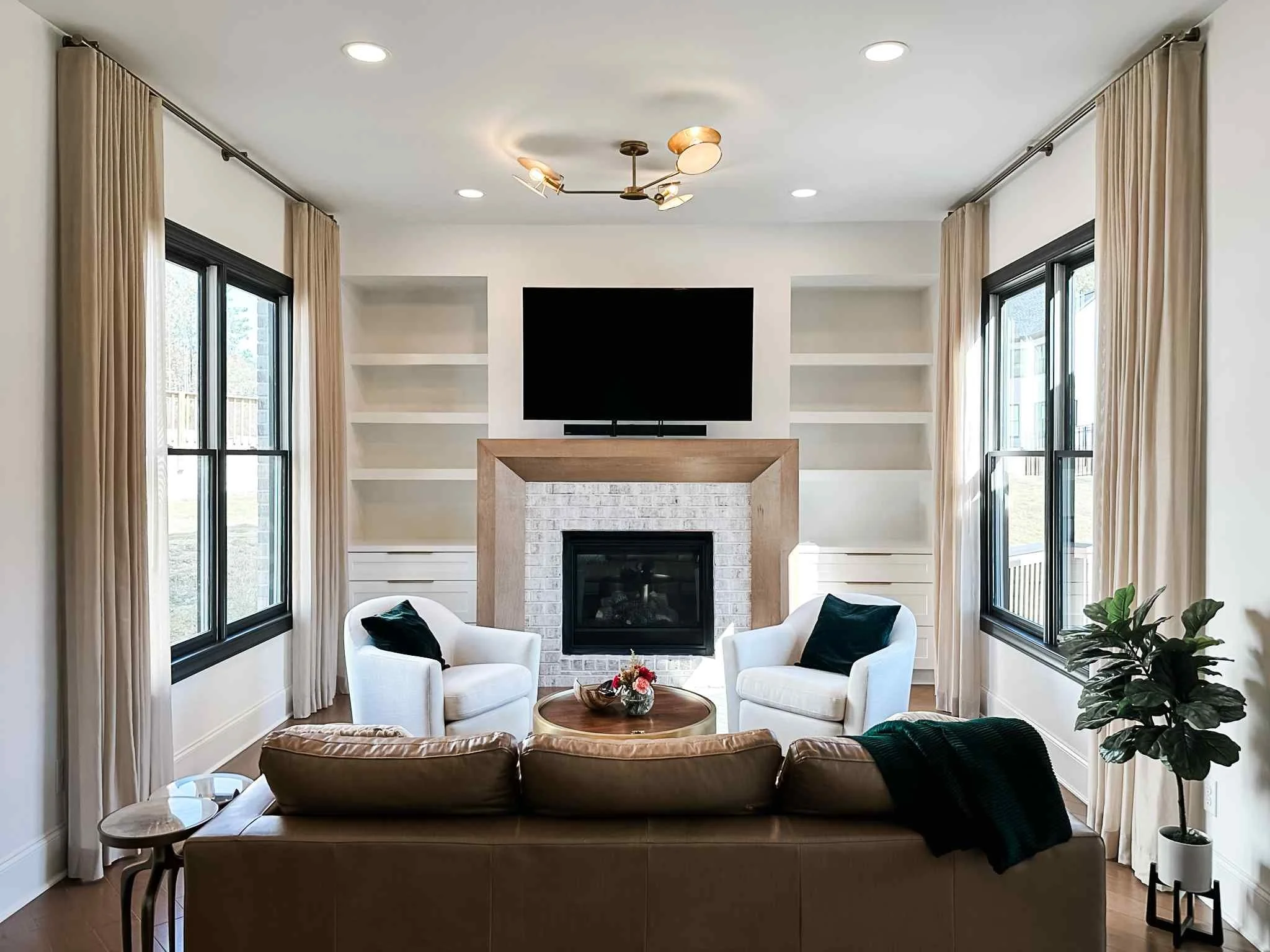 Symmetrical living room design with cream pinch pleat drapes, wood fireplace surround, and built-in shelving by Luxe Style Studio