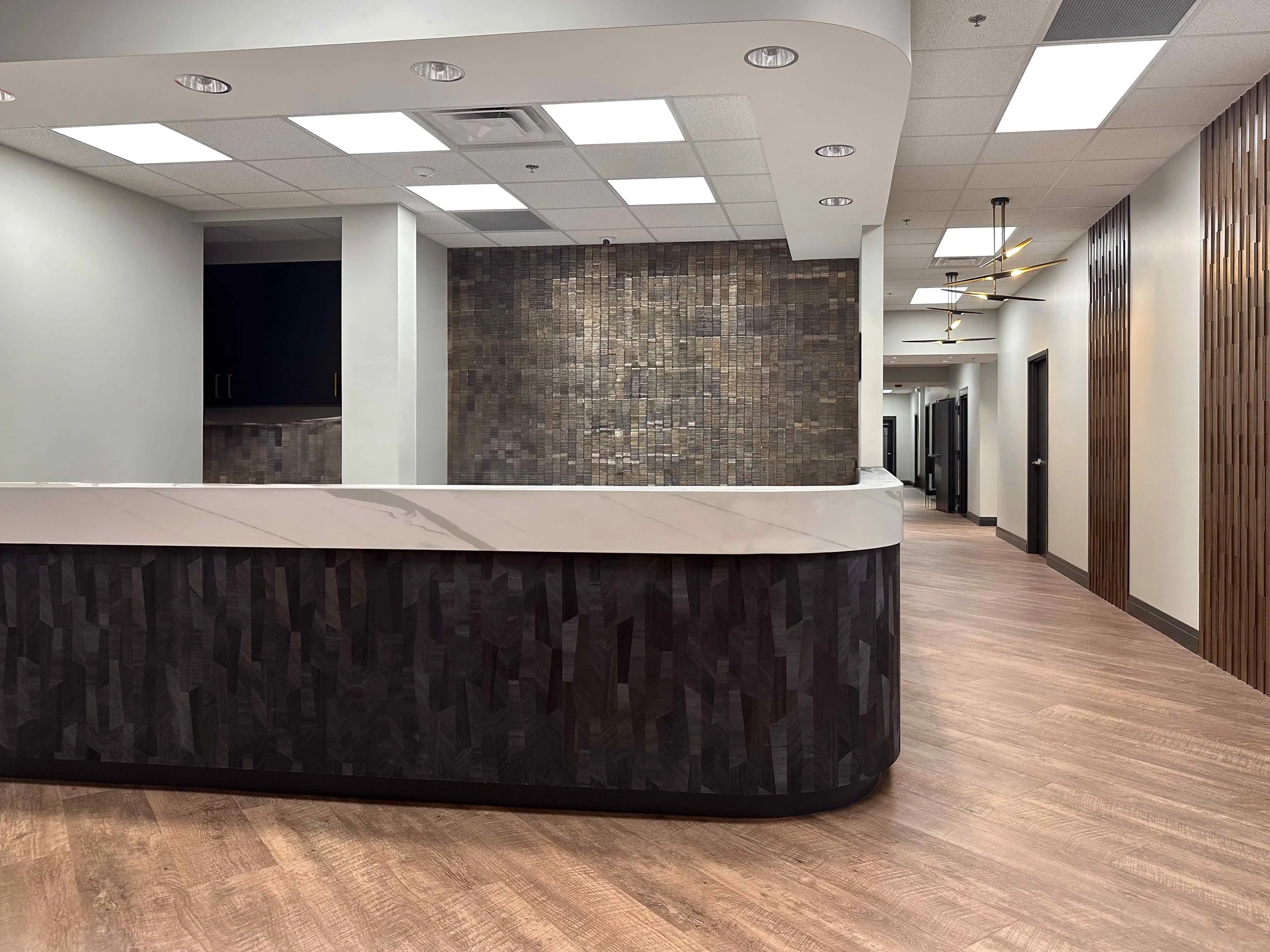 Corporate office reception desk with curved dark mosaic tile panel, white marble countertop, wood slat accent wall, brass sputnik pendant light, and wood plank floors leading to office corridor, interior design by Luxe Style Studio
