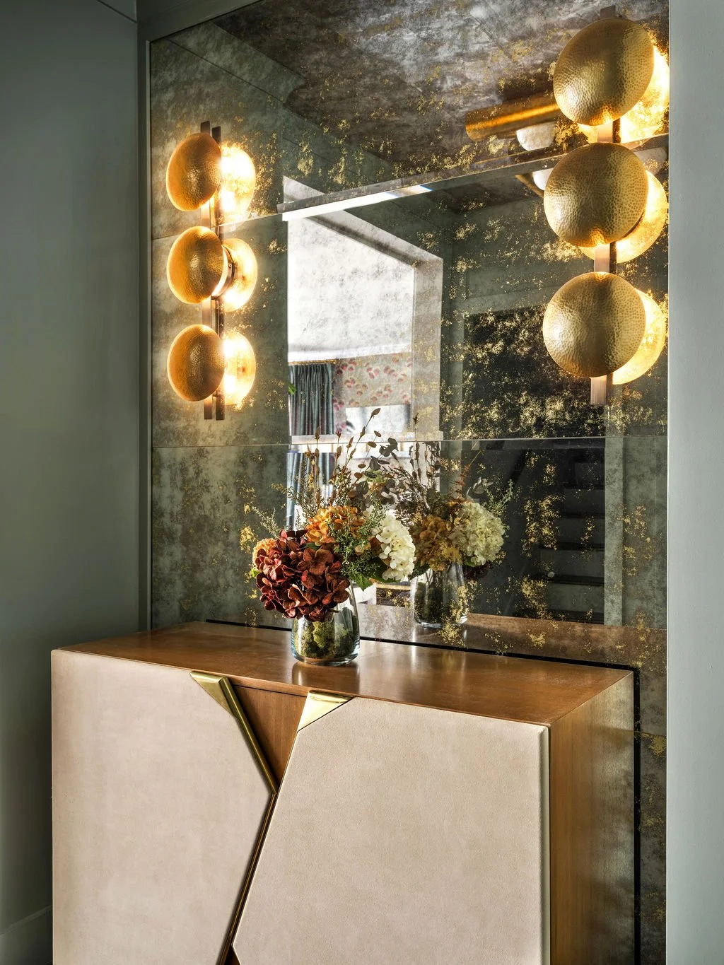 Glamorous vanity vignette with full antiqued mirror wall, hammered brass dome sconces, geometric leather and brass console, and lush floral arrangement, interior design by Luxe Style Studio