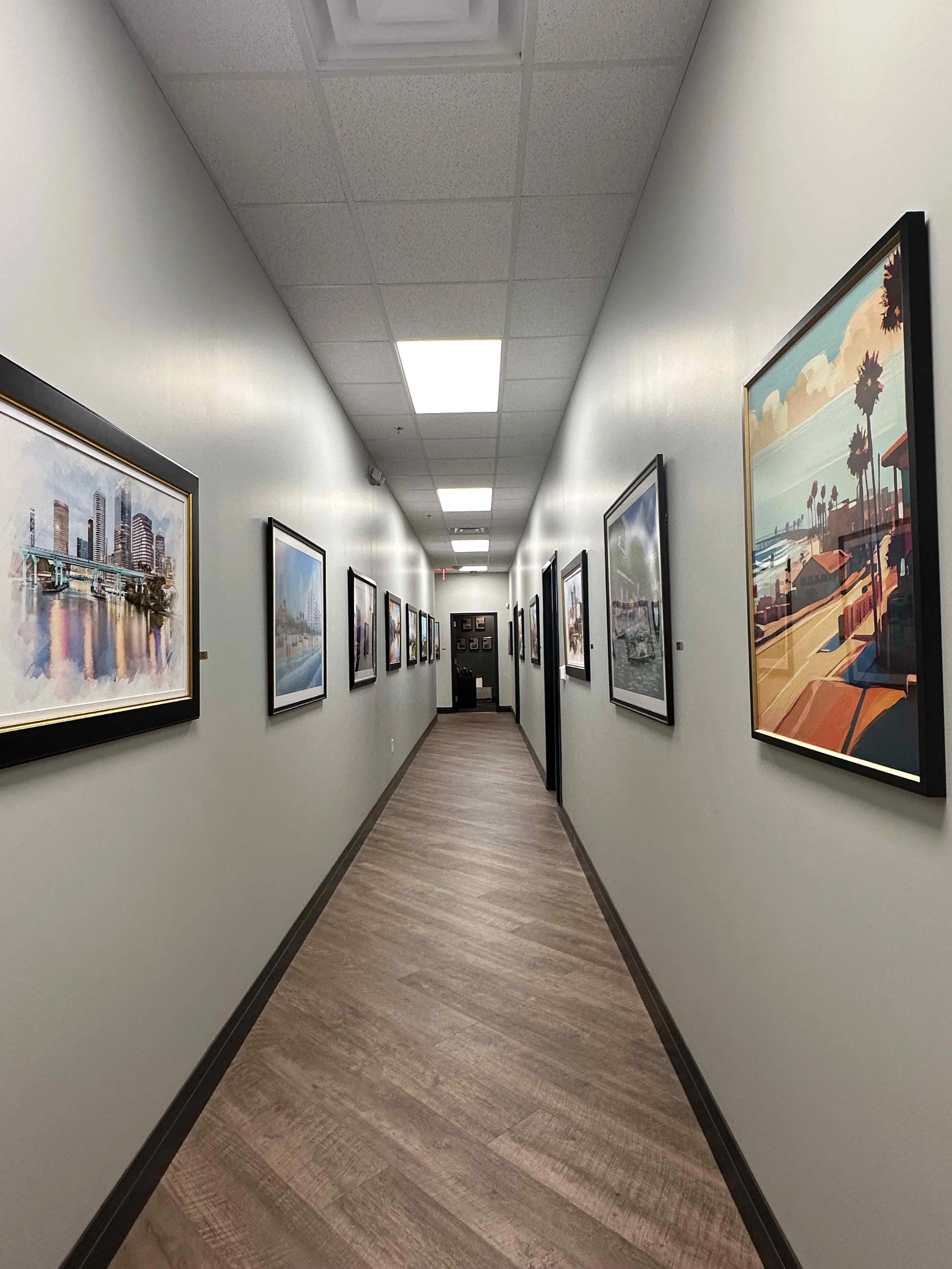 Long corporate office hallway with bilateral gallery wall of large-scale cityscape and travel artwork in black frames, wood plank floors, and recessed panel ceiling, interior design by Luxe Style Studio