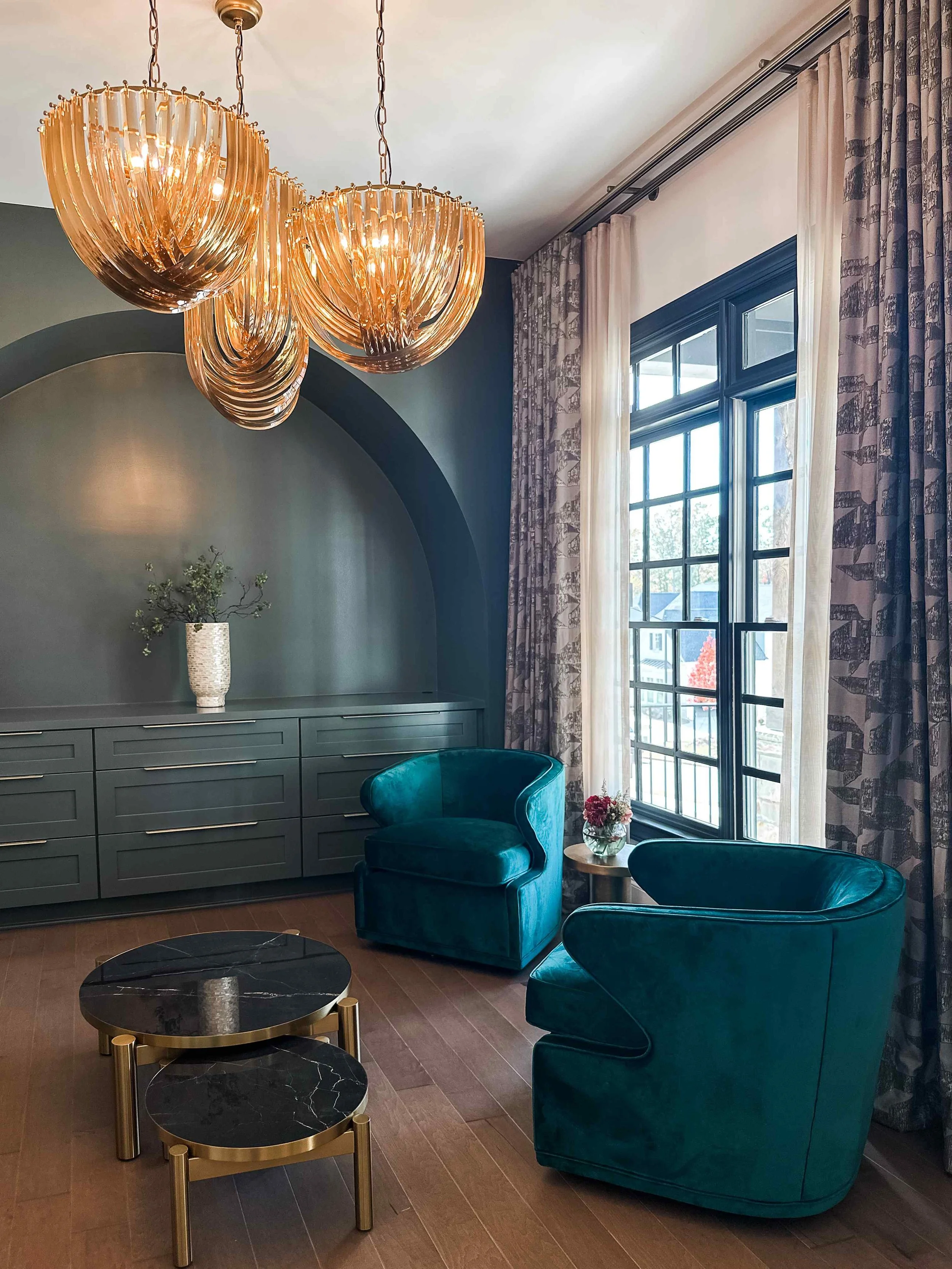 Moody jewel-toned sitting room with teal velvet barrel chairs, arched wall niche, and amber glass chandelier pair by Luxe Style Studio