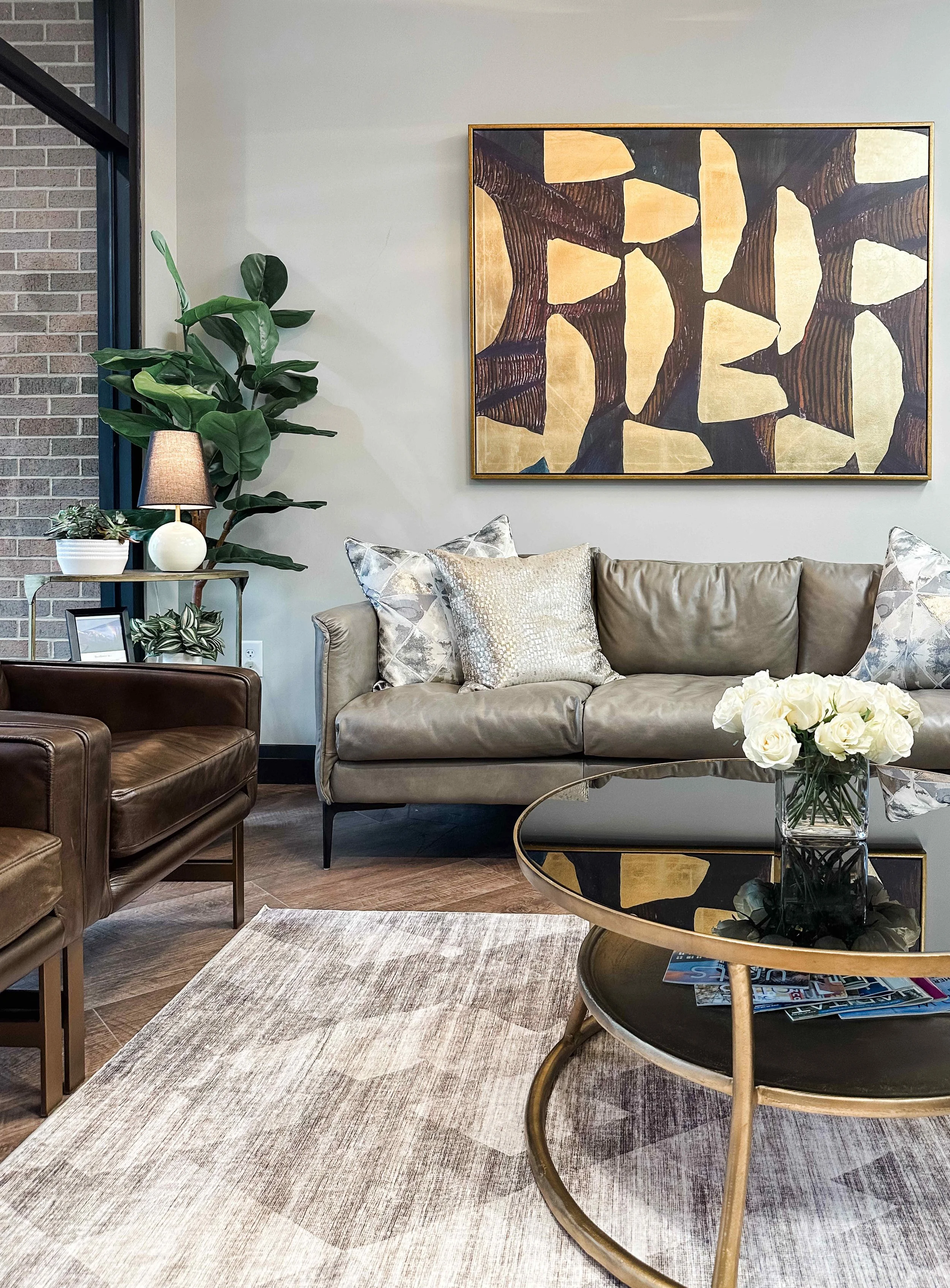 Corporate office reception lounge with taupe leather sofa, metallic pillows, gold leaf abstract artwork, glass and brass coffee table with white roses, dark leather club chair, fiddle leaf fig tree, and exposed brick wall with black steel windows, in