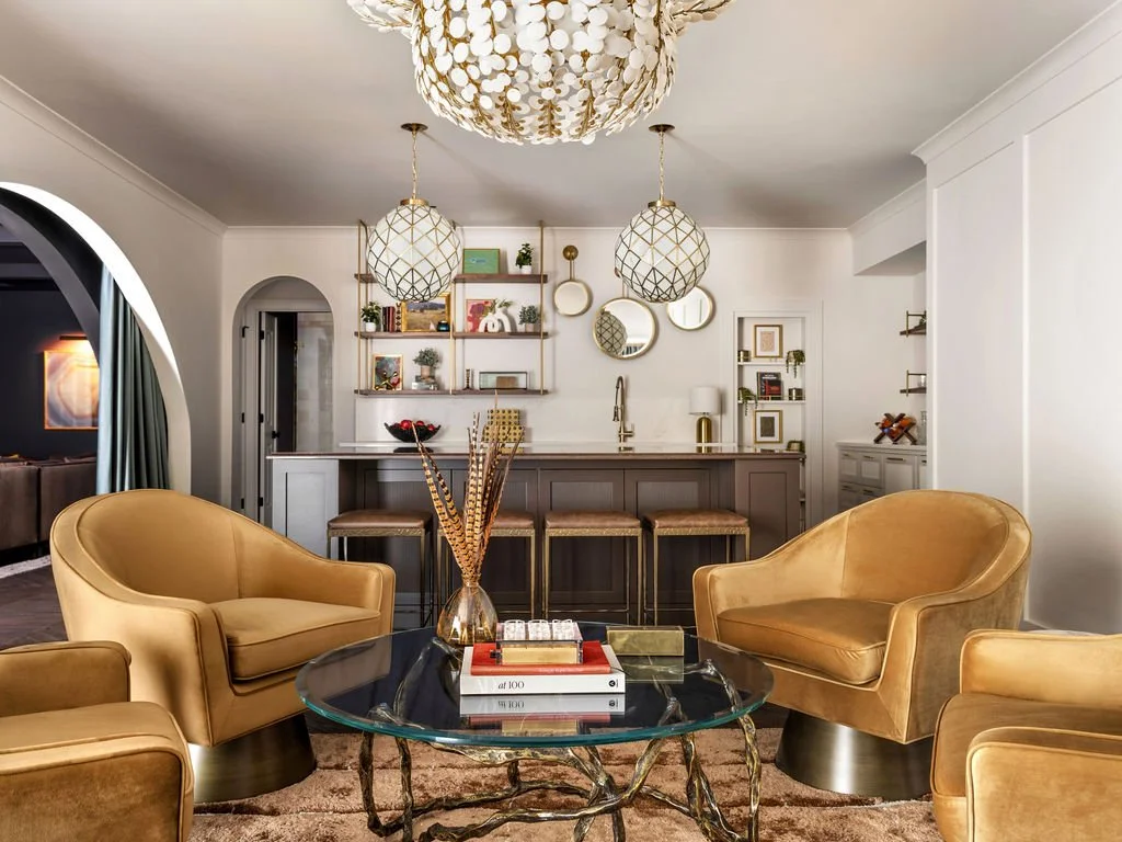 Glamorous living room with gold velvet swivel chairs, sculptural brass and glass coffee table, capiz shell chandelier, and open view to kitchen bar, interior design by Luxe Style Studio