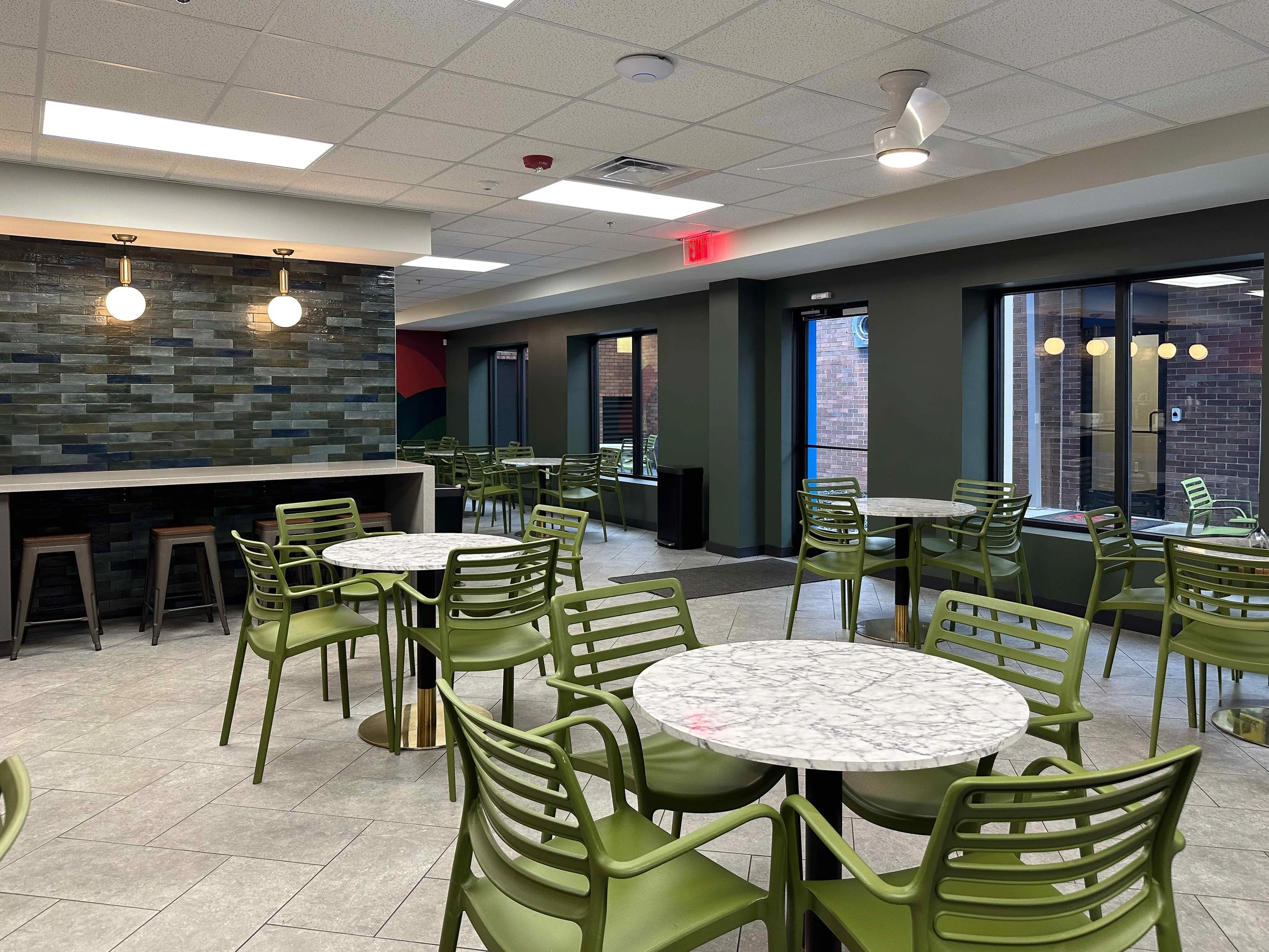 Large corporate break room café seating area with sage green bistro chairs, round marble top tables, multicolor zellige tile accent wall, brass globe pendants, and floor-to-ceiling black steel windows, interior design by Luxe Style Studio