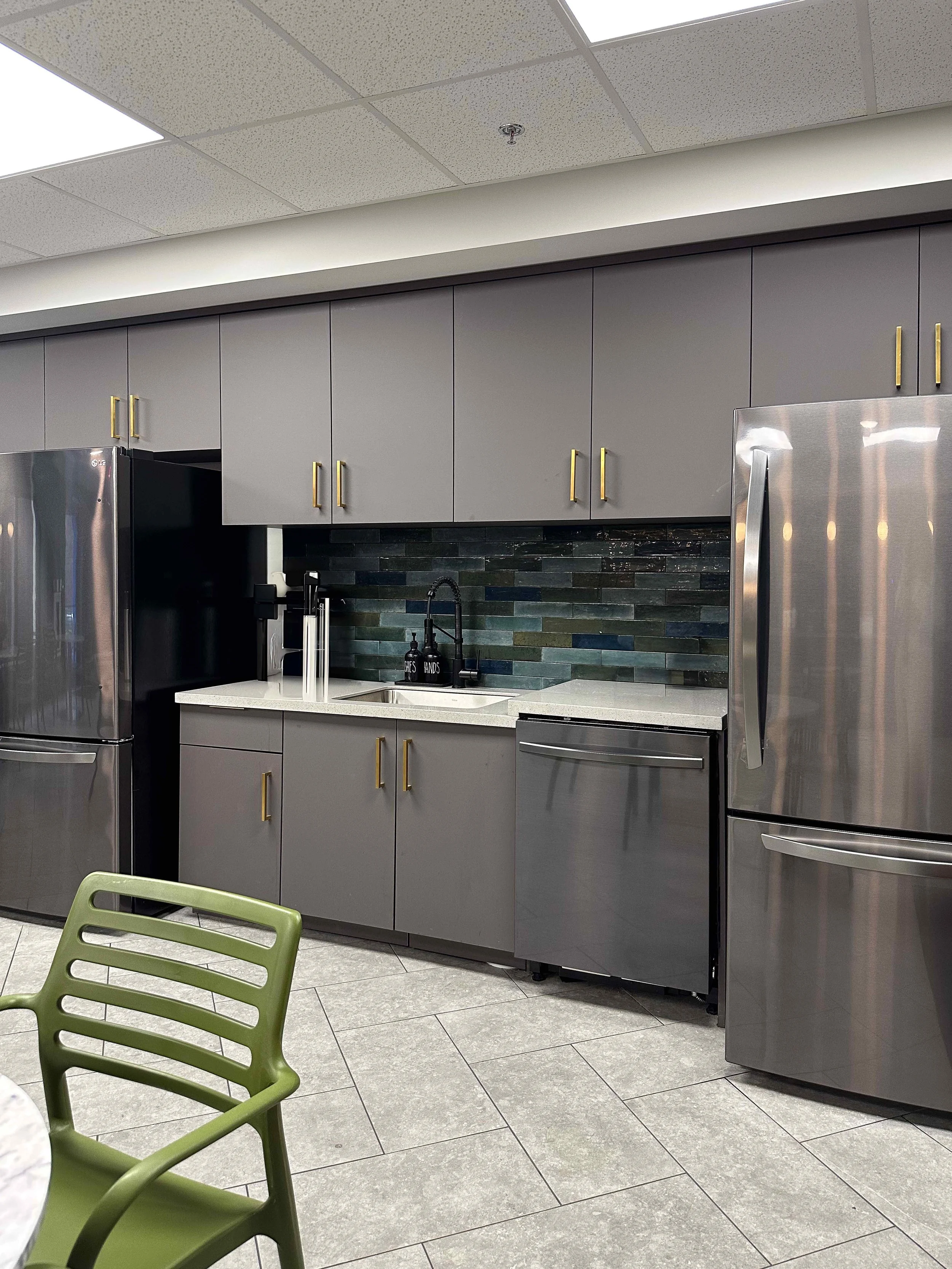 Corporate break room kitchen with floor-to-ceiling gray flat-front cabinetry, brass bar pull hardware, multicolor zellige tile backsplash, quartz countertop, and stainless steel appliances, interior design by Luxe Style Studio