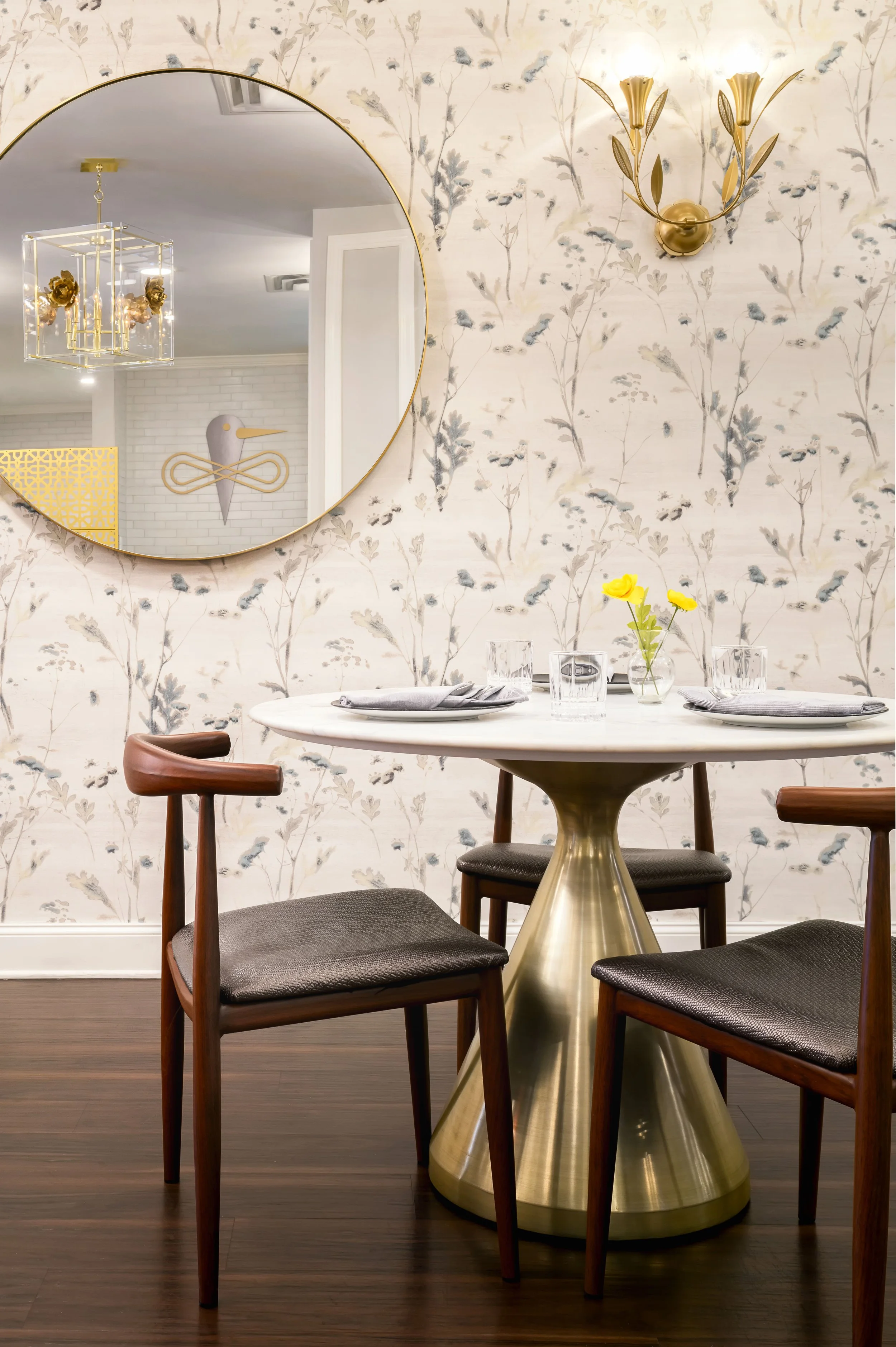 Zinnia Café & Market dining vignette with a marble and brass table, round brass mirror, gold tulip sconce, and botanical wallpaper, designed by Ashley Schneider
