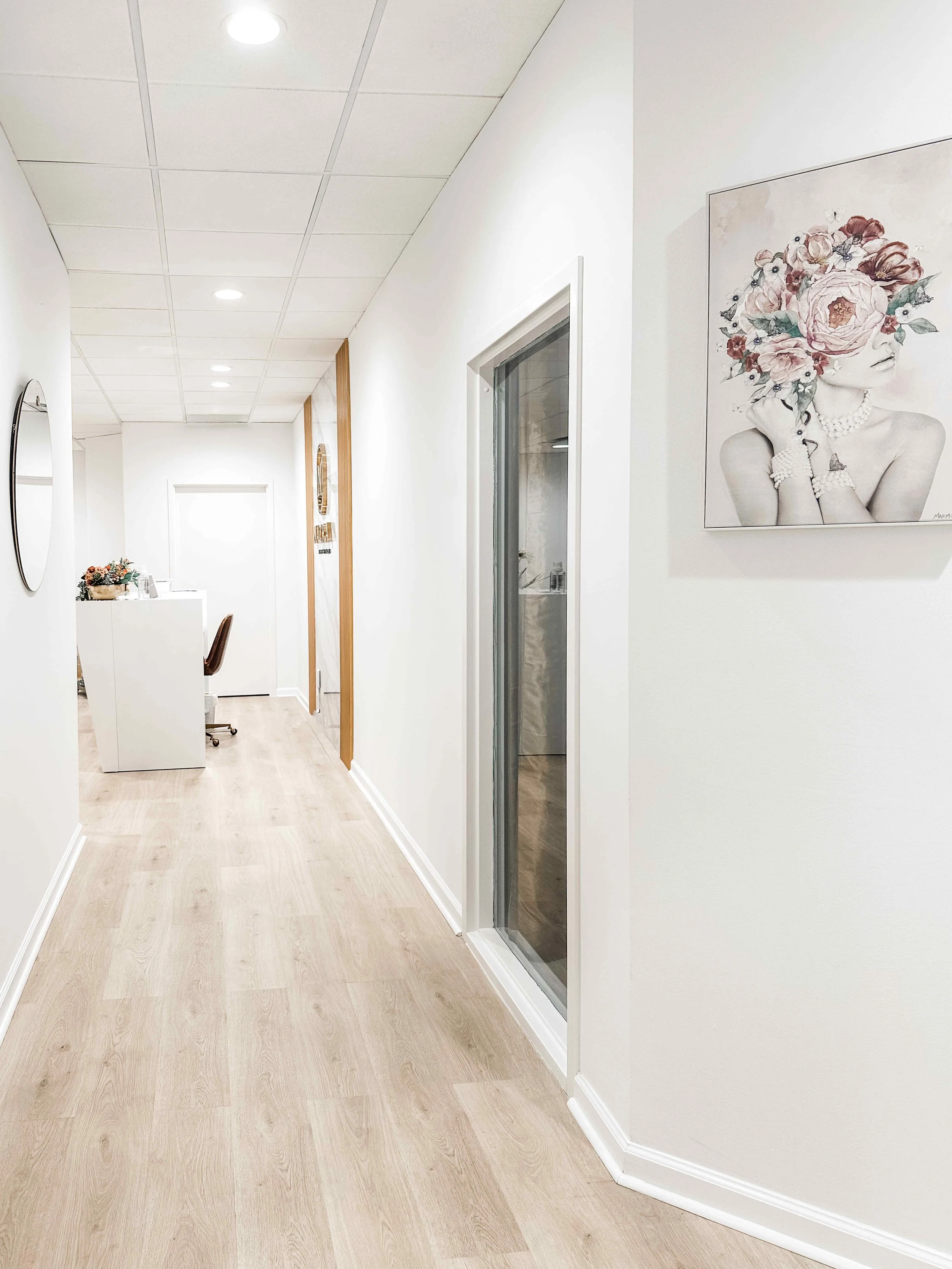 Bright clinical hallway in luxury medical spa with white oak floors, clean white walls, framed floral artwork, and glass-paneled treatment room doors leading to reception, interior design by Luxe Style Studio