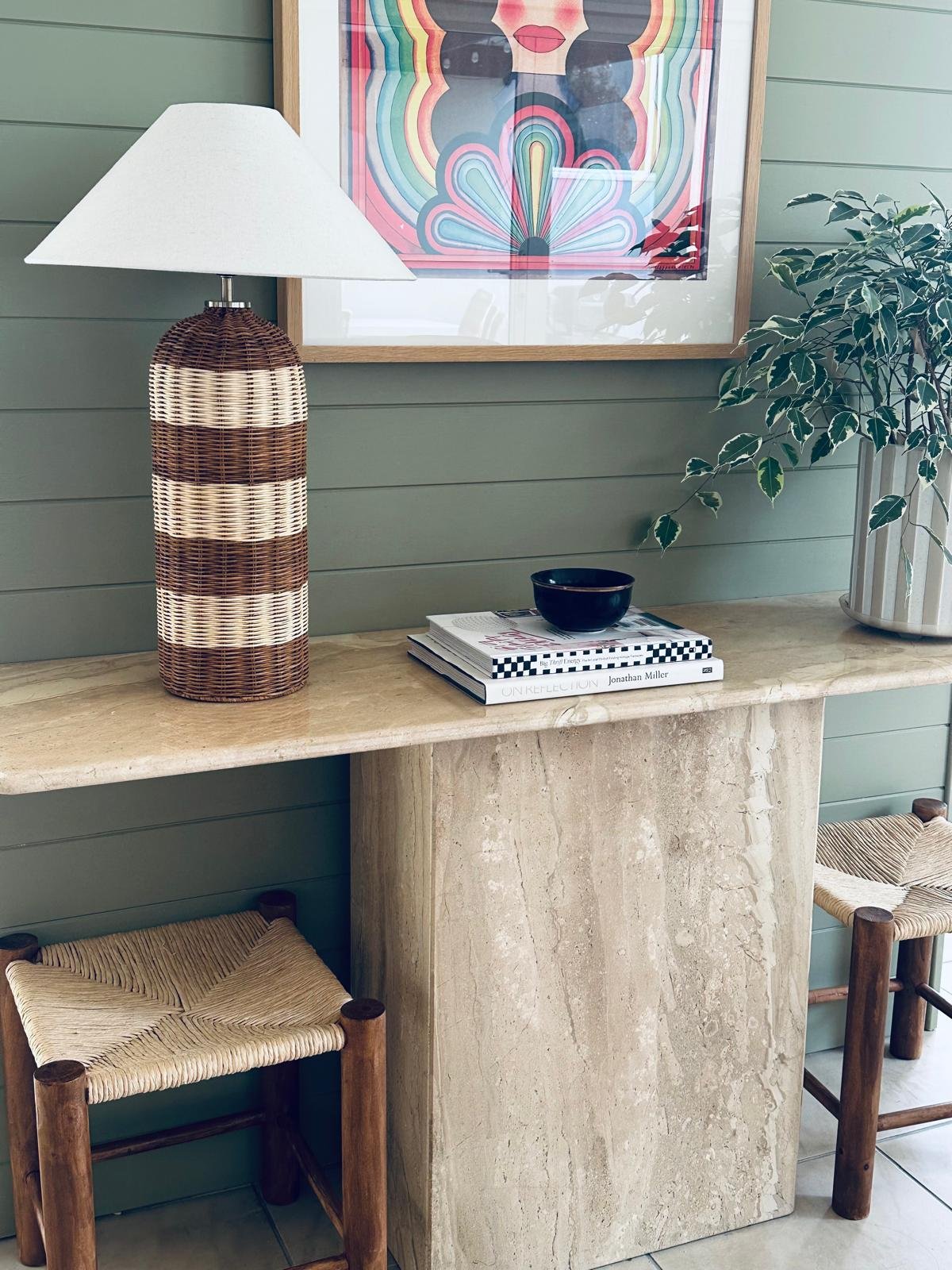 Decorative indoor scene with a wicker table lamp, a wall print of a stylized woman, a black bowl, a stack of books, and two wooden stools with woven seats against a green paneled wall.