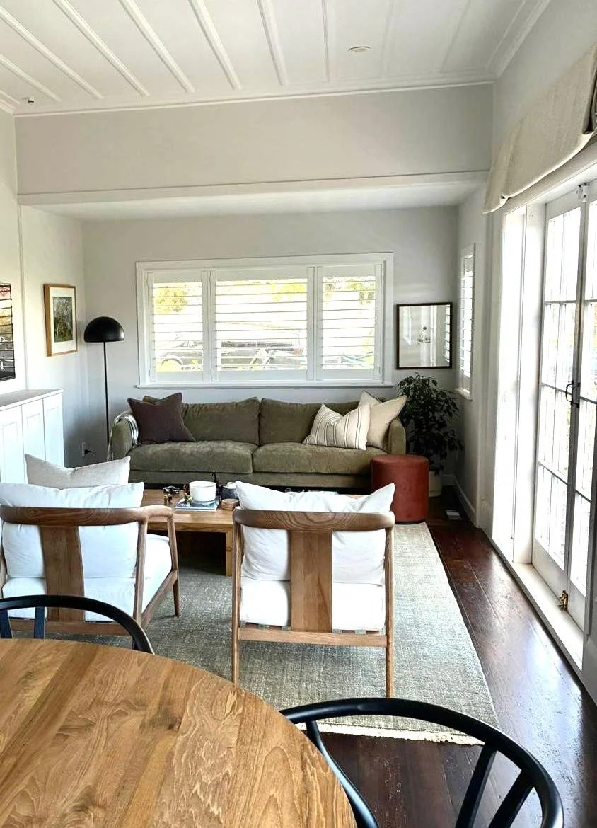 Living room with a green sofa, white pillows, black floor lamp, framed artwork, window with white blinds, potted plant, ottoman, beige rug, French doors, wooden dining table with black and wooden chairs.