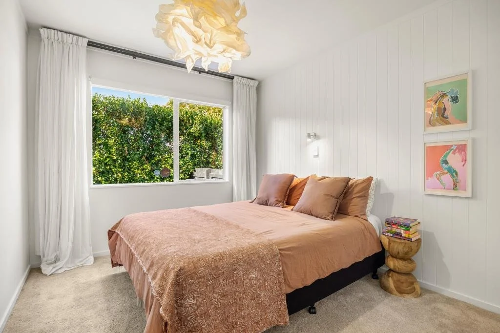 A cozy bedroom with beige bedding, a nightstand with books, colorful wall art, a large window with white curtains, and a modern light fixture hanging from the ceiling.