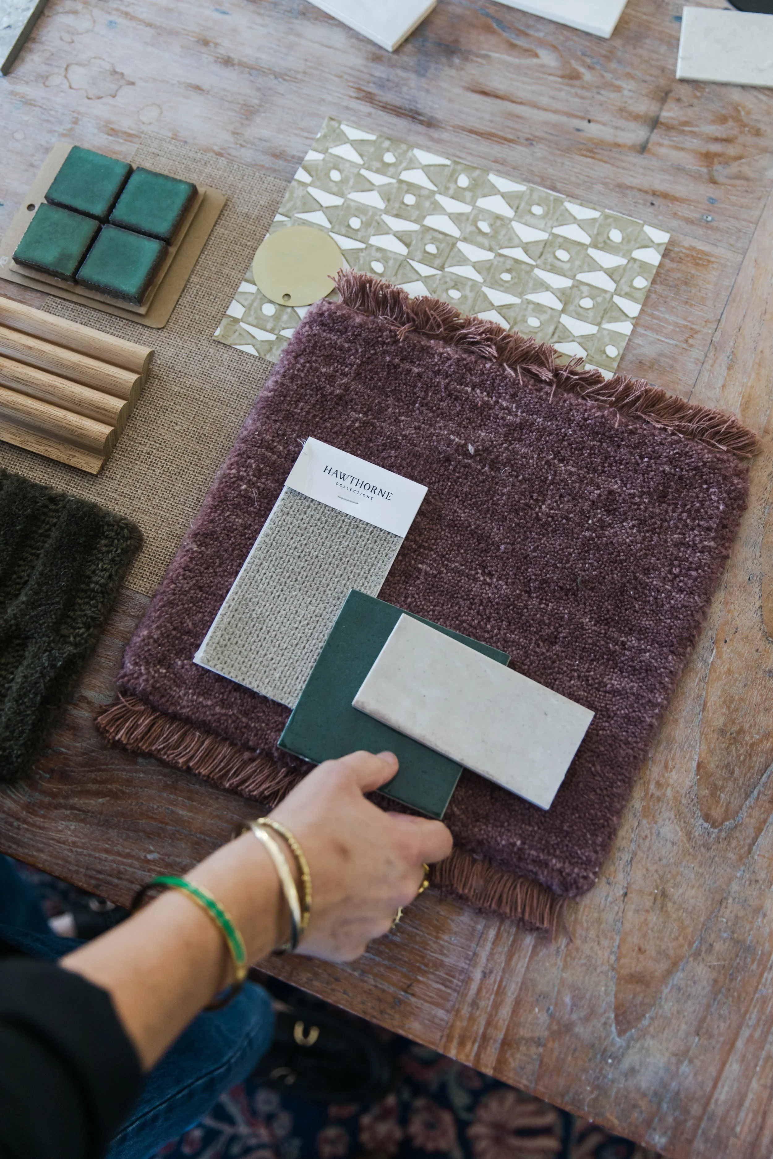 Person holding green tile sample over a fabric swatch on a wooden table surrounded by other tile samples, a tile sample book, and small tiles.