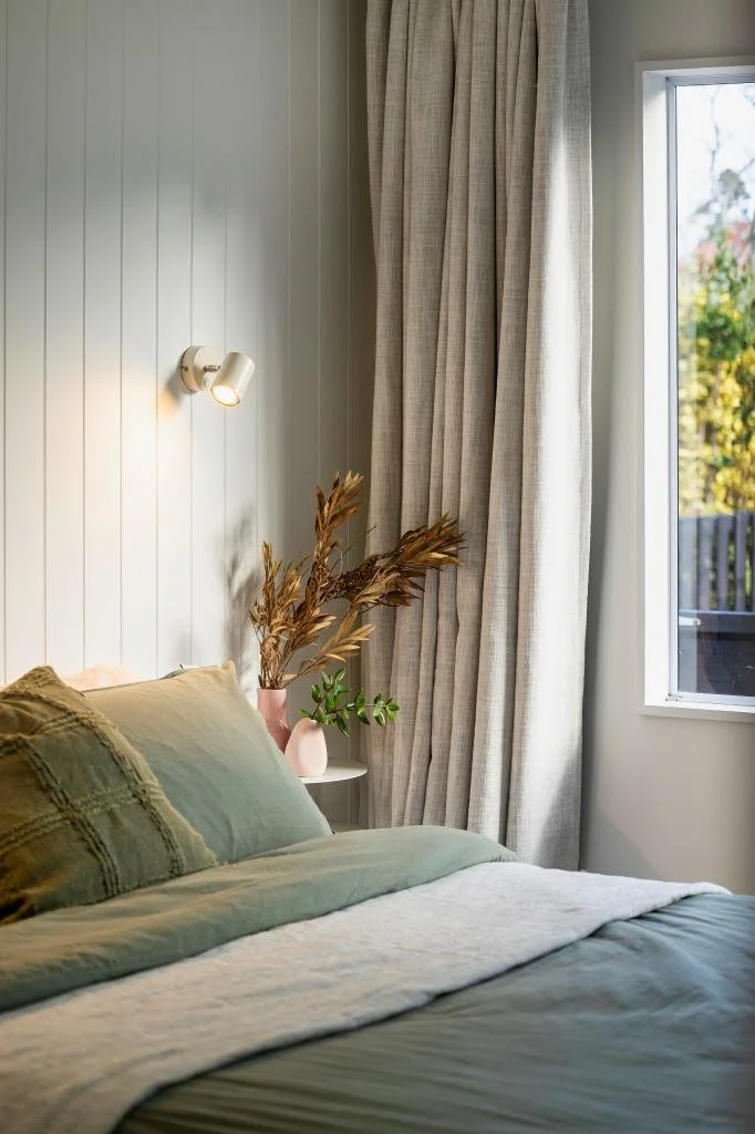 A cozy bedroom corner with a bed featuring green and beige bedding, a pink vase with dried brown leaves, a green plant in a pink pot, beige curtains, and a window showing a scenic outdoor view.