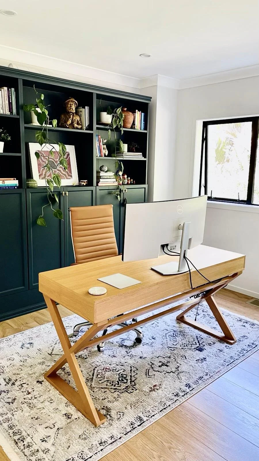 Home office with wooden desk, beige office chair, large white monitor, patterned rug, black bookshelf with books and plants, white walls, and a window with natural light.