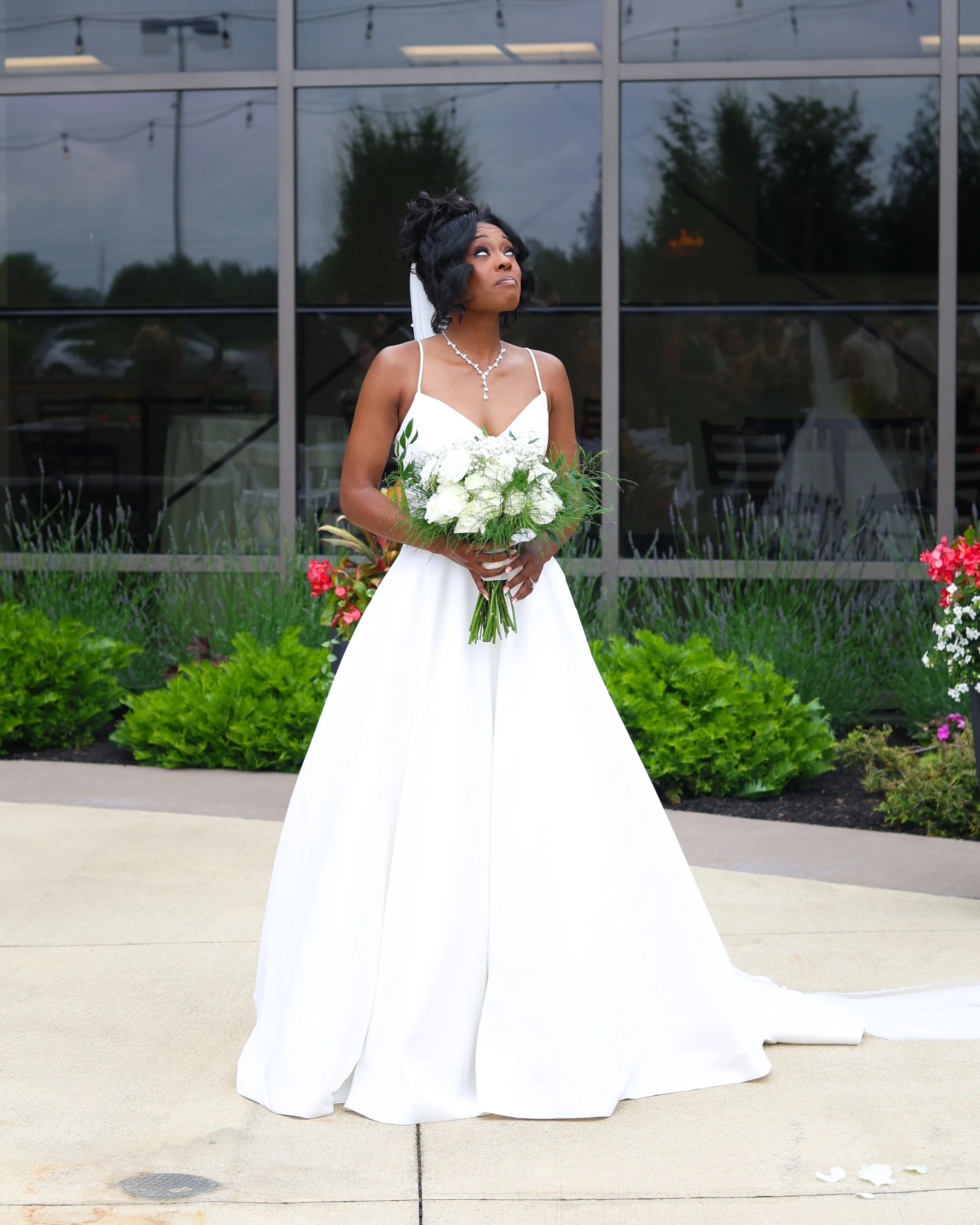 A bride in a white wedding dress holding a bouquet of white flowers, standing outdoors in front of a building with large glass windows and greenery.