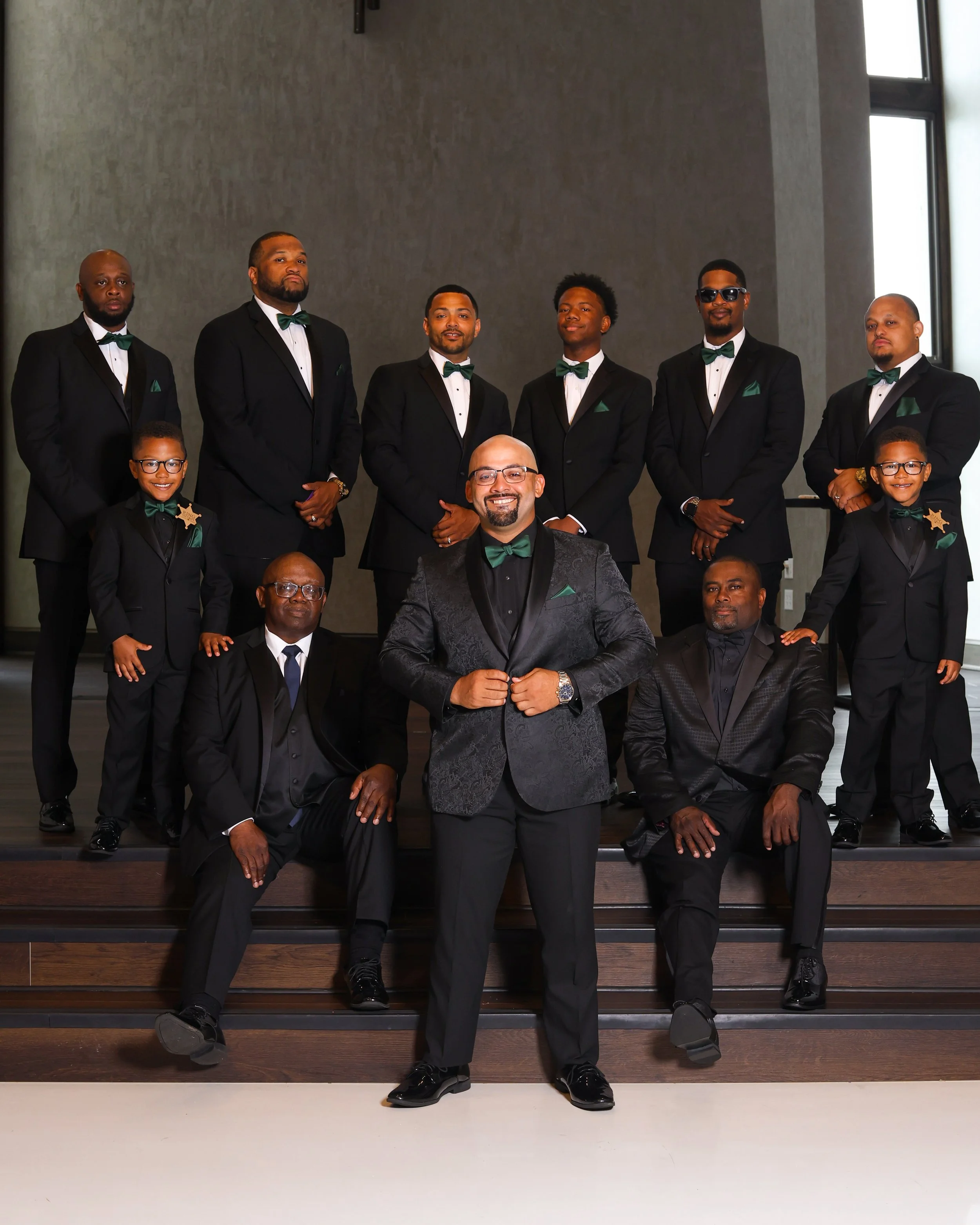 A group of eleven men and boys dressed in black tuxedos with green bow ties and pocket squares, posing together on wooden steps in a formal setting.