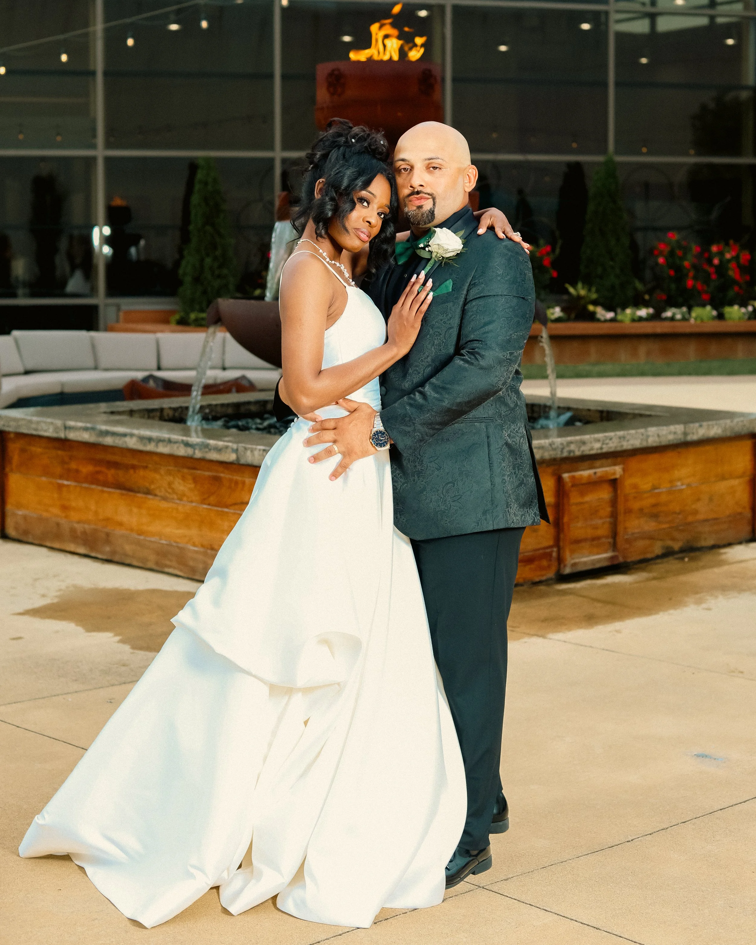 A newlywed couple posing together at their wedding reception outdoors. The bride is wearing a white wedding gown, and the groom is in a dark suit with a green bow tie and boutonniere. They are standing close in front of a fountain with a fire sculpture on top, with decorative plants and glass windows in the background.