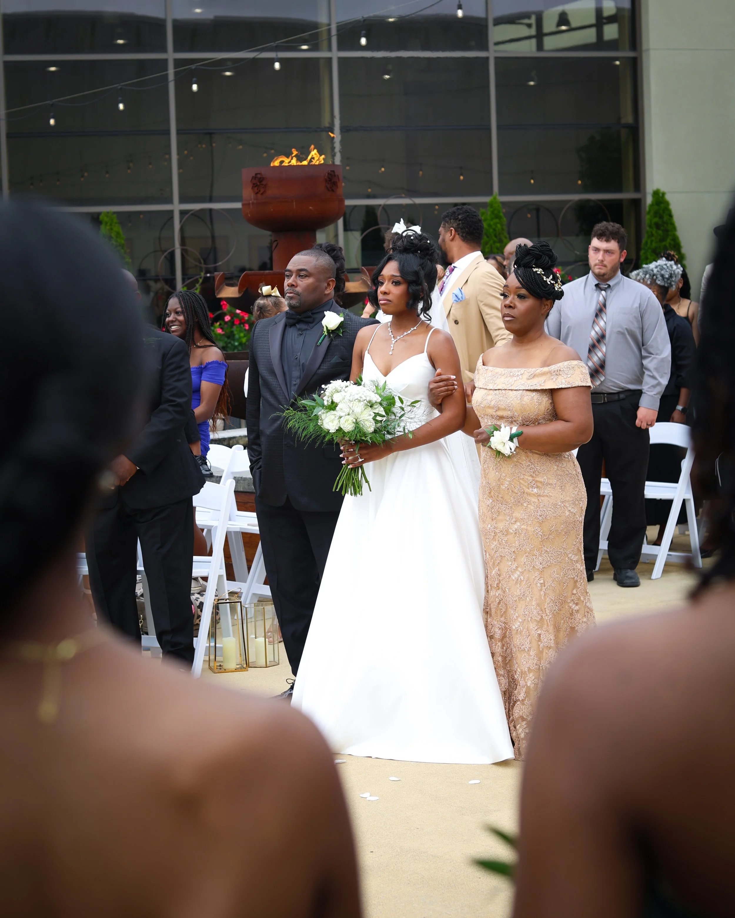 A bride in a white wedding gown holding a bouquet of greenery and white flowers, walking down the aisle with her parents at an outdoor wedding ceremony, surrounded by guests seated on white chairs.
