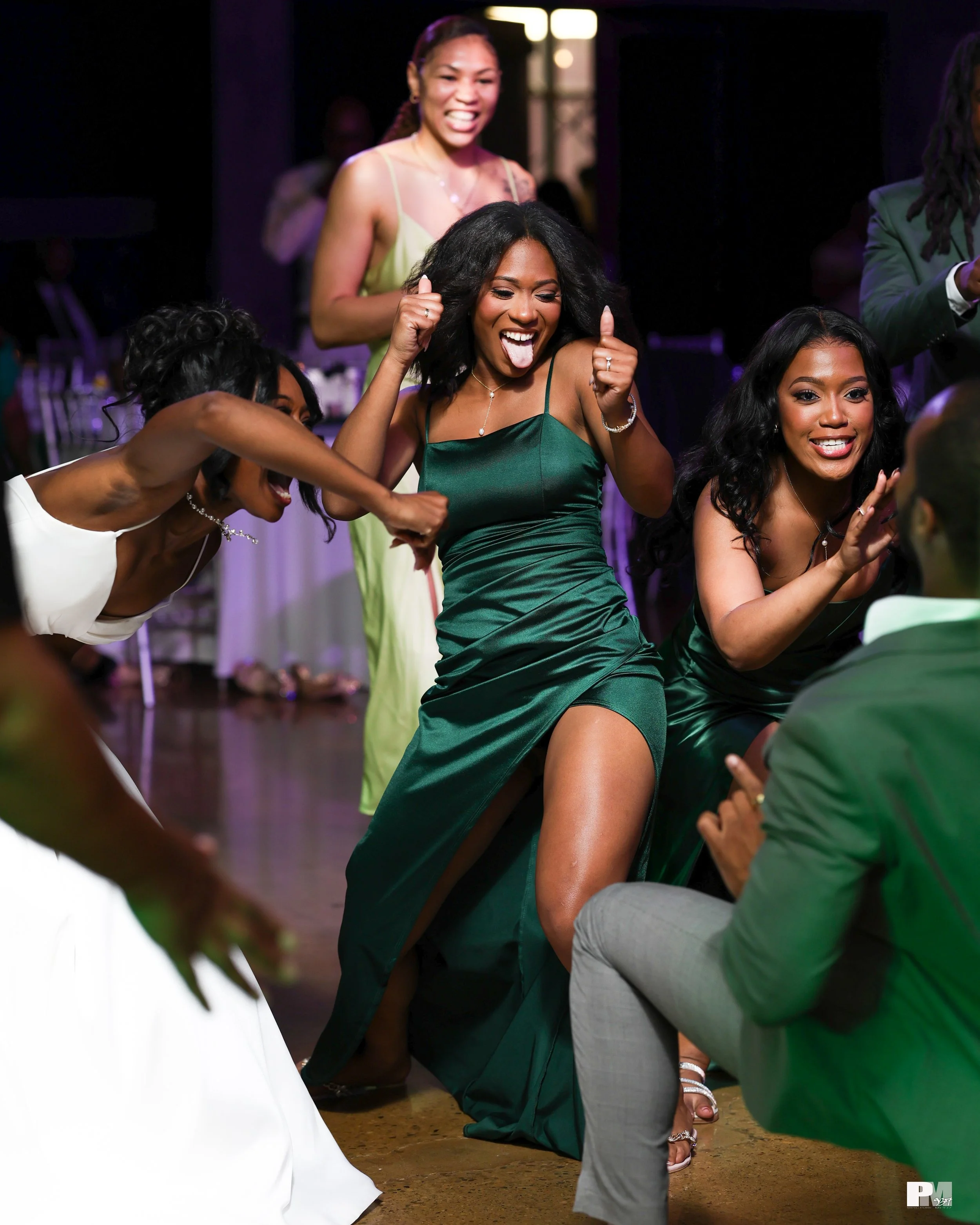 A group of women dancing and celebrating at a party or celebration, with a woman in a green satin dress prominently in the center making a happy face and dancing.