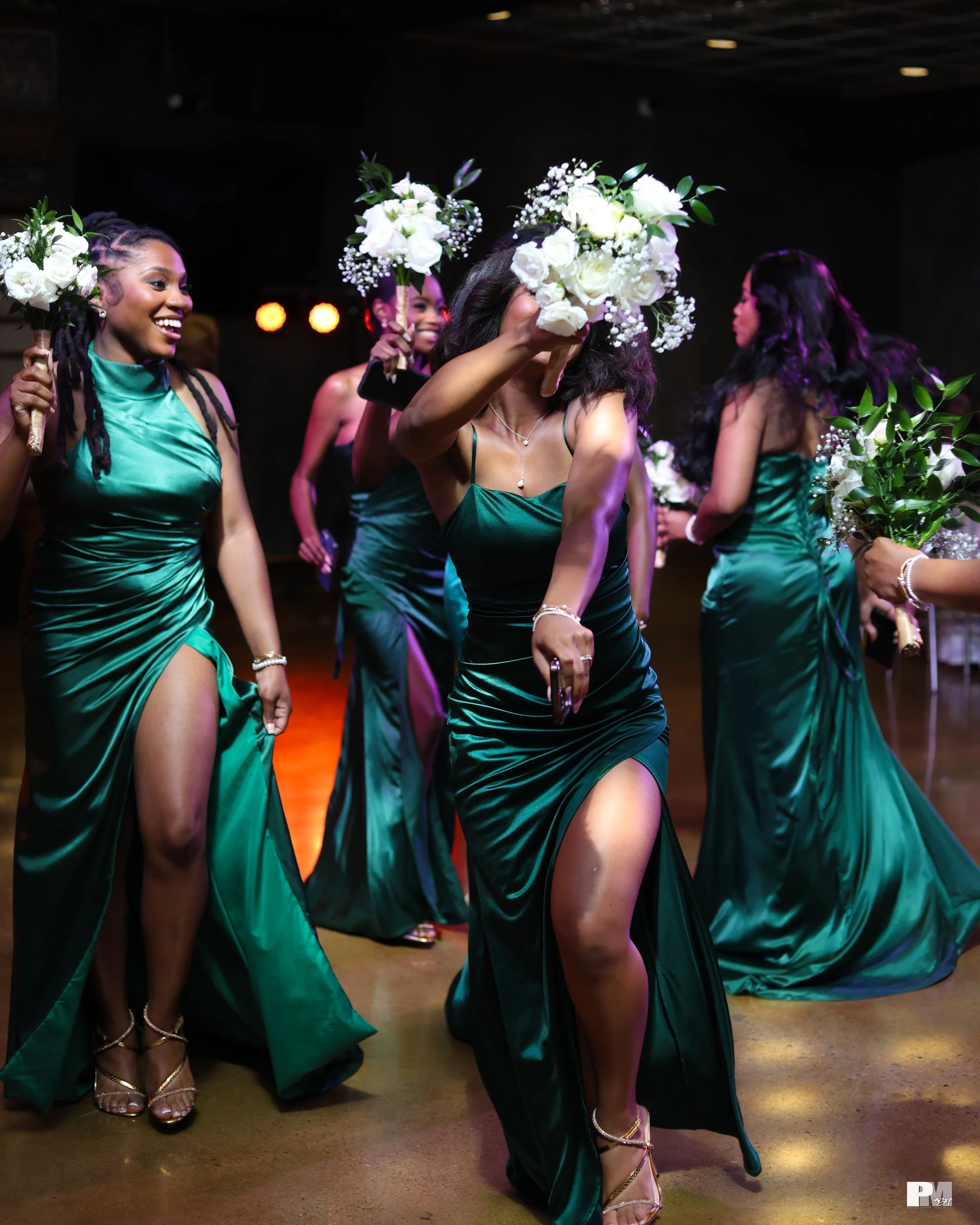 A group of women in emerald green dresses dancing and holding bouquets of white flowers at a celebration event.