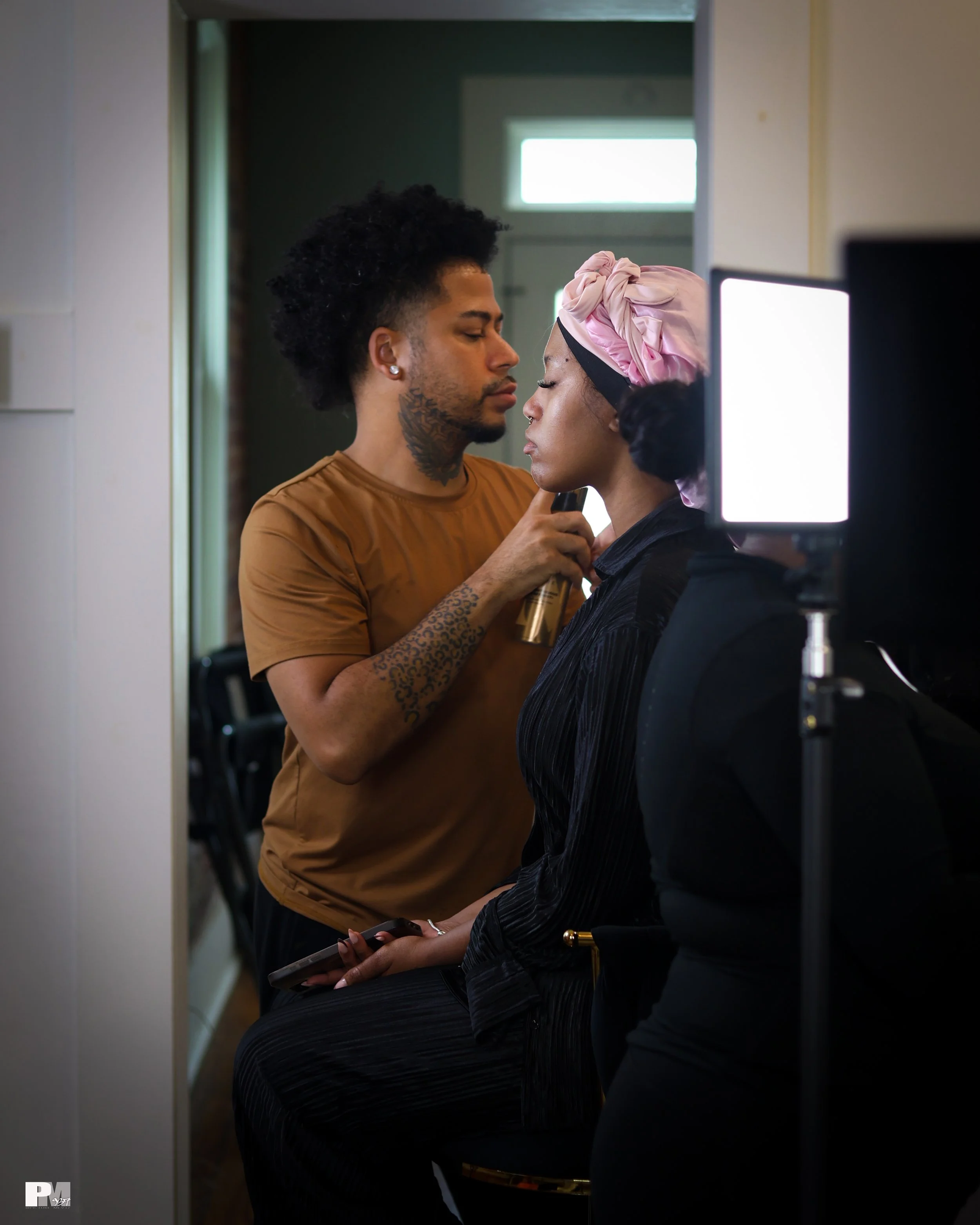 A makeup artist applying makeup to a woman sitting with her eyes closed, wearing a pink headscarf, in a well-lit room.