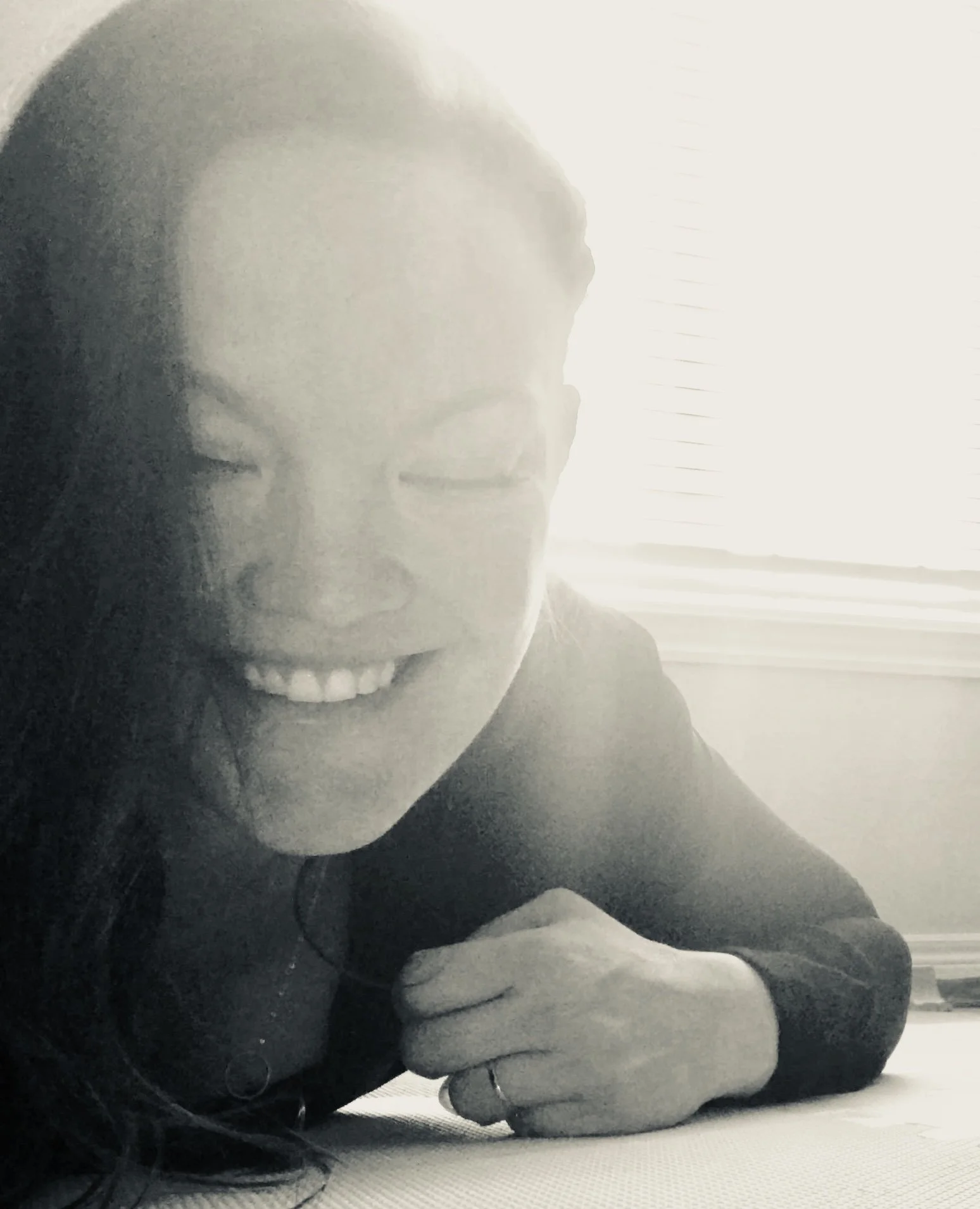 A woman with long dark hair smiling, lying on her side on a light-colored surface, wearing a dark top and a ring on her finger.