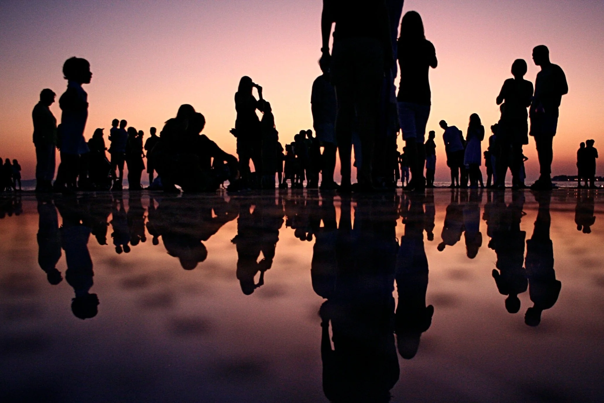Silhouettes of people on a beach at sunset, with their reflections visible in the wet sand.