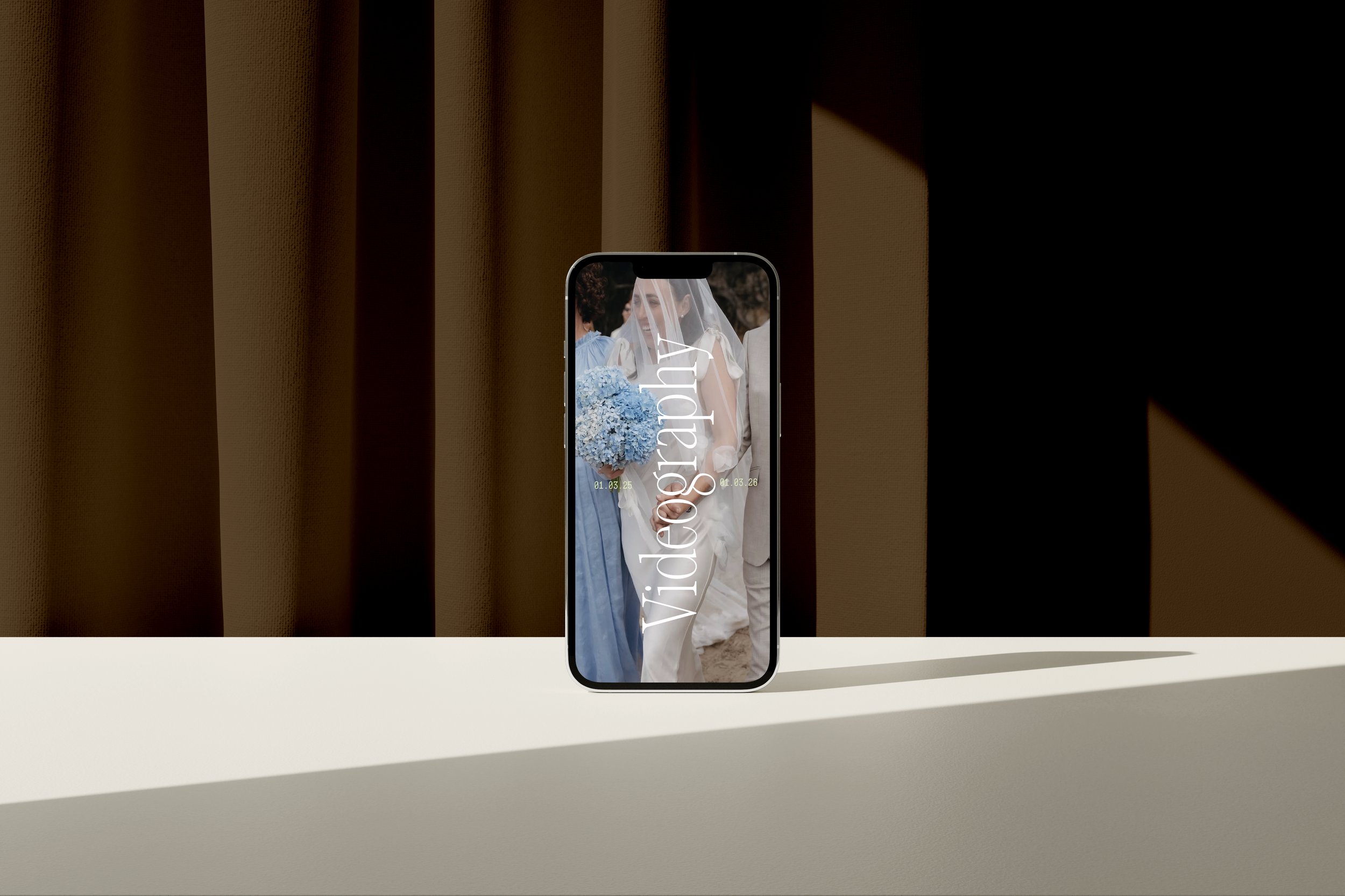 A smartphone standing on a white surface displays a wedding photo with the word "videography" overlaid, featuring a bride holding a bouquet of blue flowers, surrounded by guests.