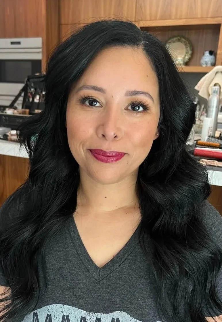 A woman with long black hair and makeup, wearing a dark gray T-shirt with the word 'MAMA' and a bear graphic, sitting in a kitchen or dining area with a wooden backsplash and countertop, and makeup items on the counter.