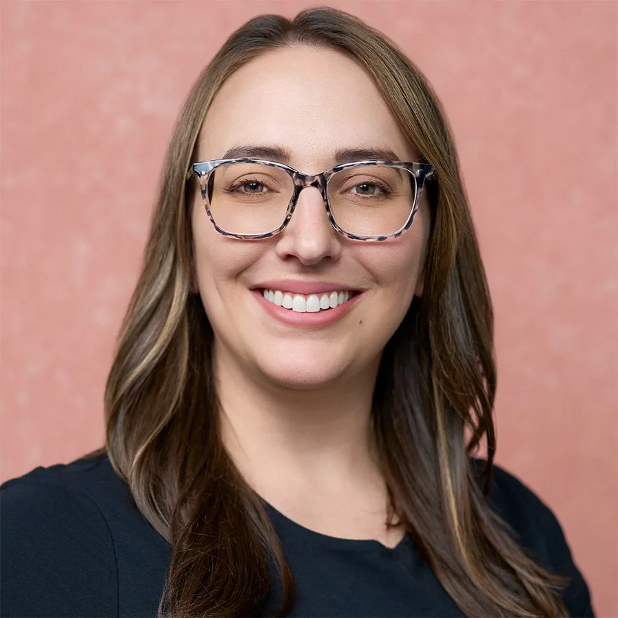 A woman with long brown hair, wearing glasses, smiling in front of a pink background.