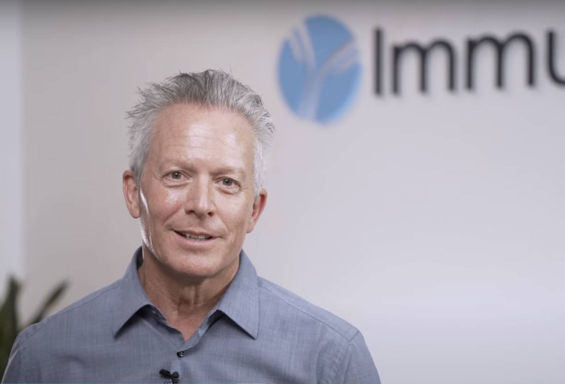 A man with gray hair and a blue shirt standing in front of a wall with a logo that says 'Immu' with a blue circular design.