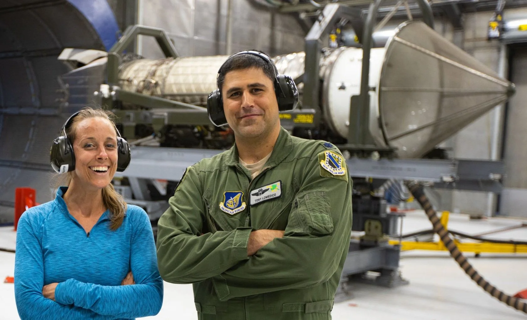 Celene Di Stasio and US Air Force Lt. Col in front of a spacecraft inside a hangar, both wearing protective headphones. The woman is smiling with arms crossed, and the man is in a military uniform with a NASA patch, looking confidently at the camera.